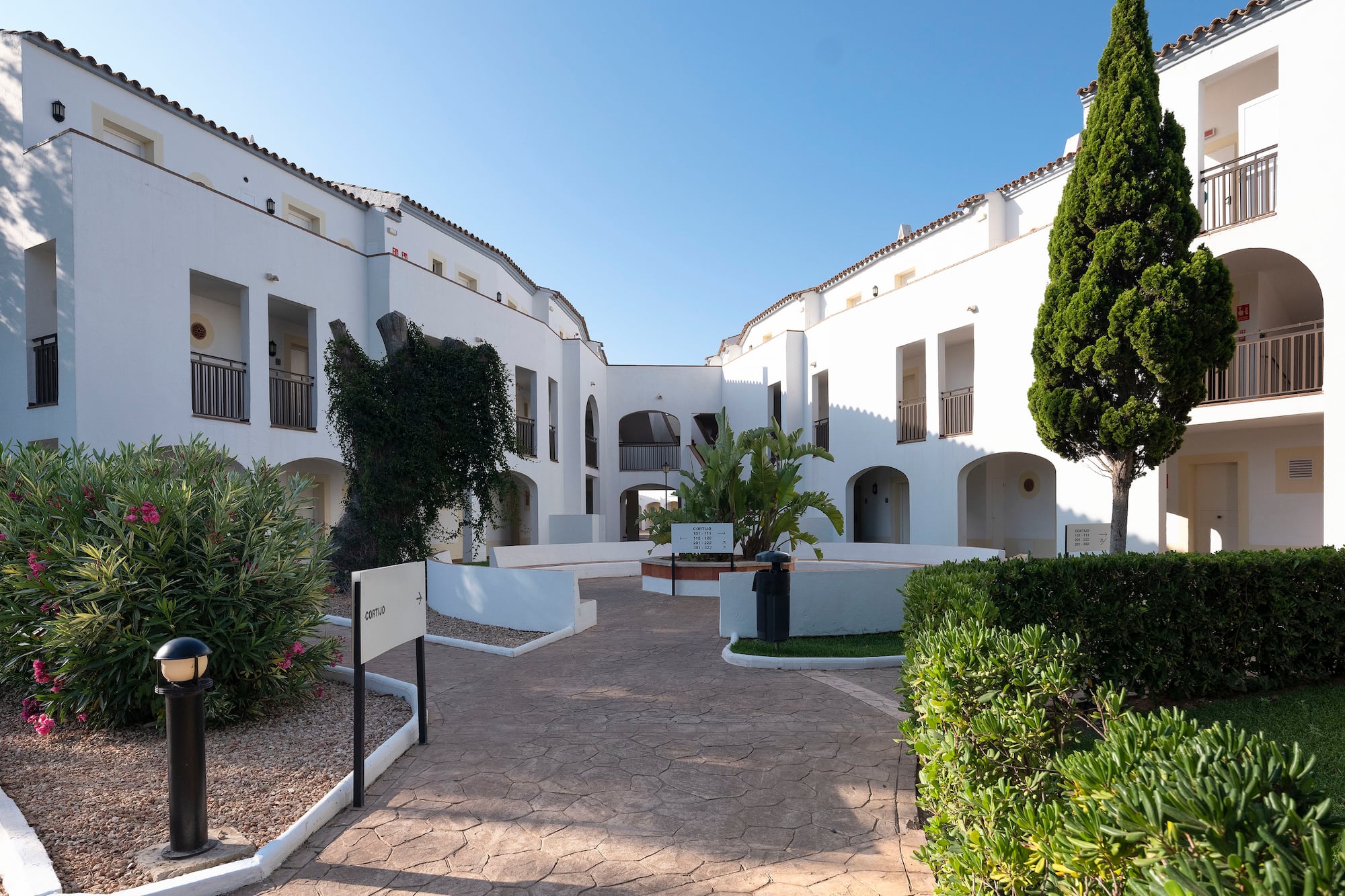 Sunny white resort courtyard with balconies, paved path, and green landscaping.