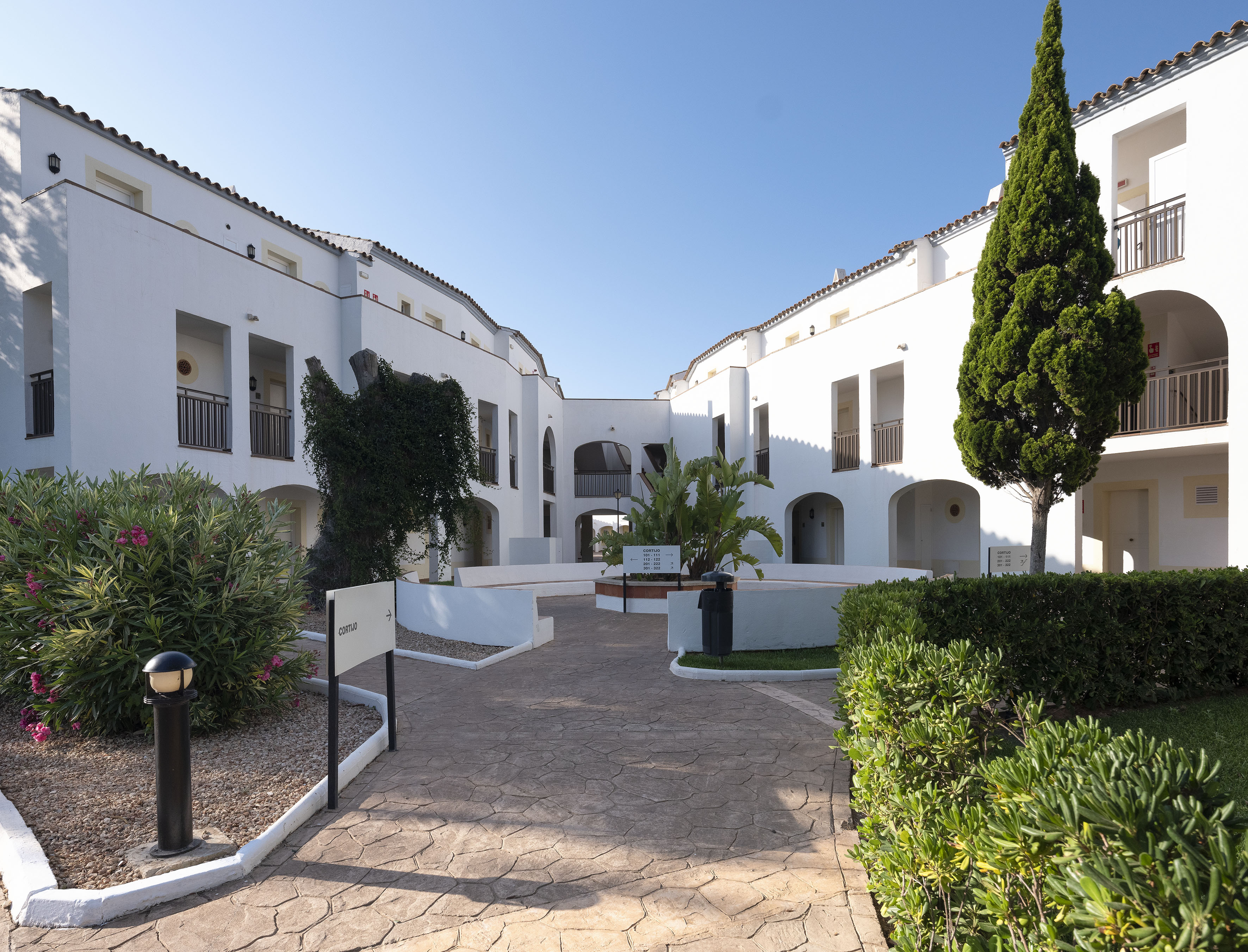 Sunny white resort courtyard with balconies, paved path, and green landscaping.