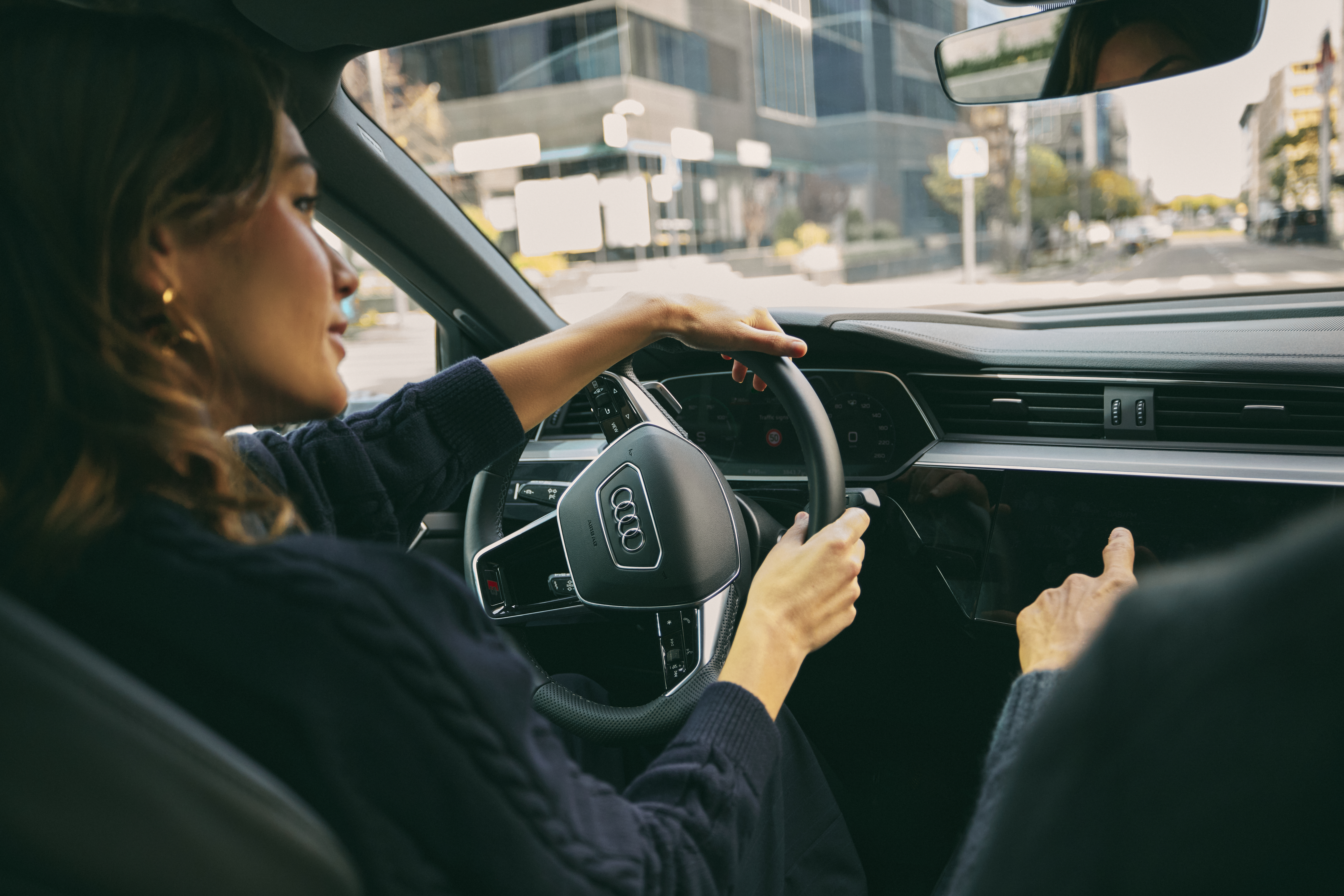 Woman driving Audi in city, hands on steering wheel and infotainment screen.