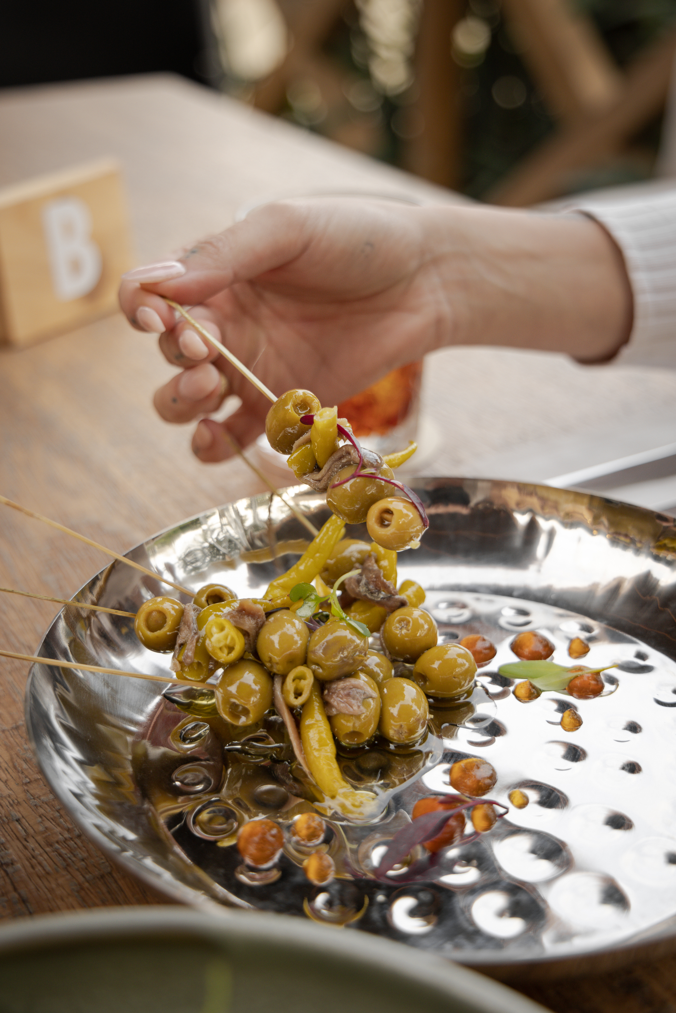 Hand lifting olive and anchovy tapas skewer with peppers from a silver plate.
