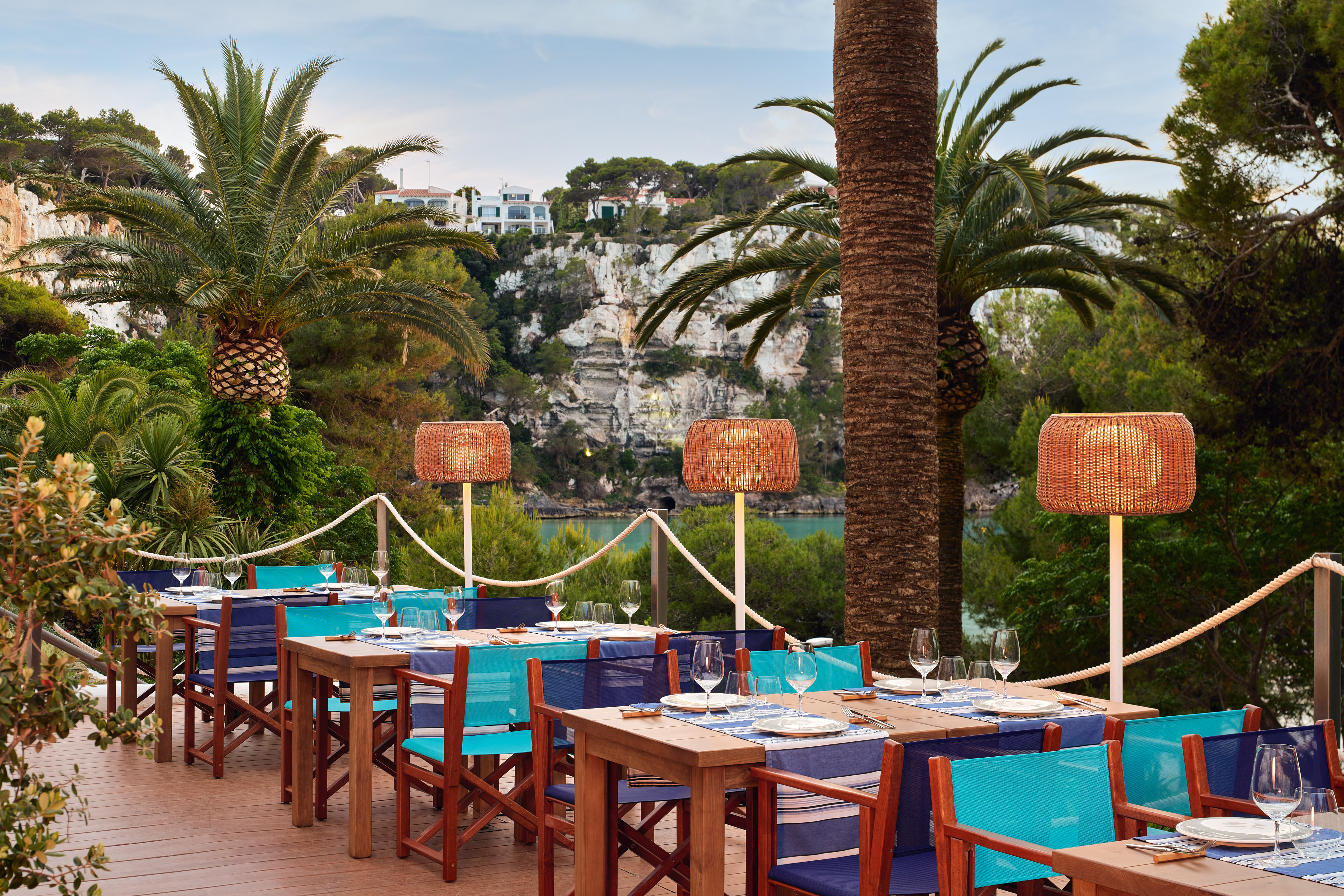 Outdoor dining terrace with set tables, palm trees, and coastal bay views.