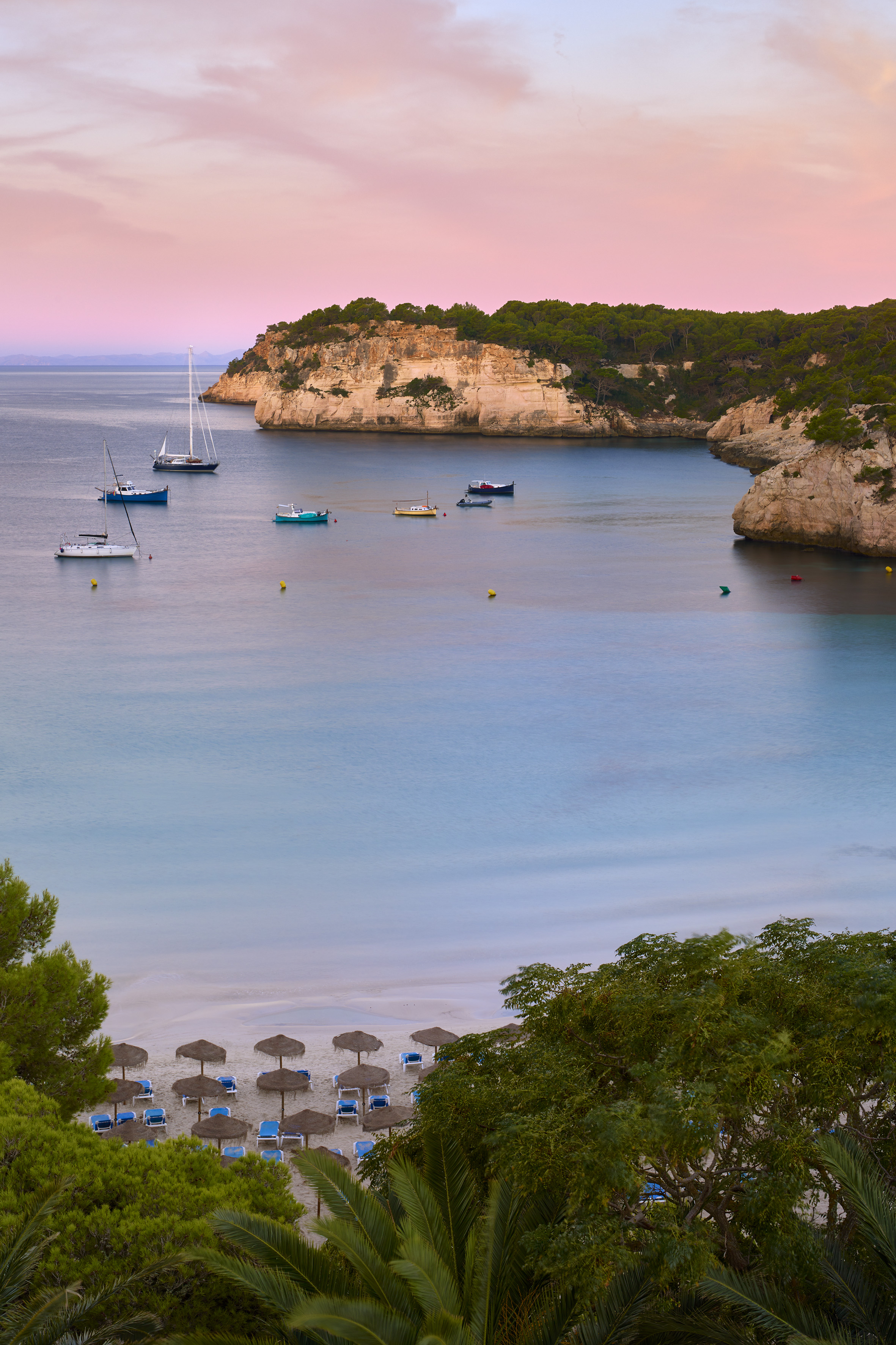 Pink sunset over tranquil bay, sailboats, sandy beach with umbrellas, cliffs.