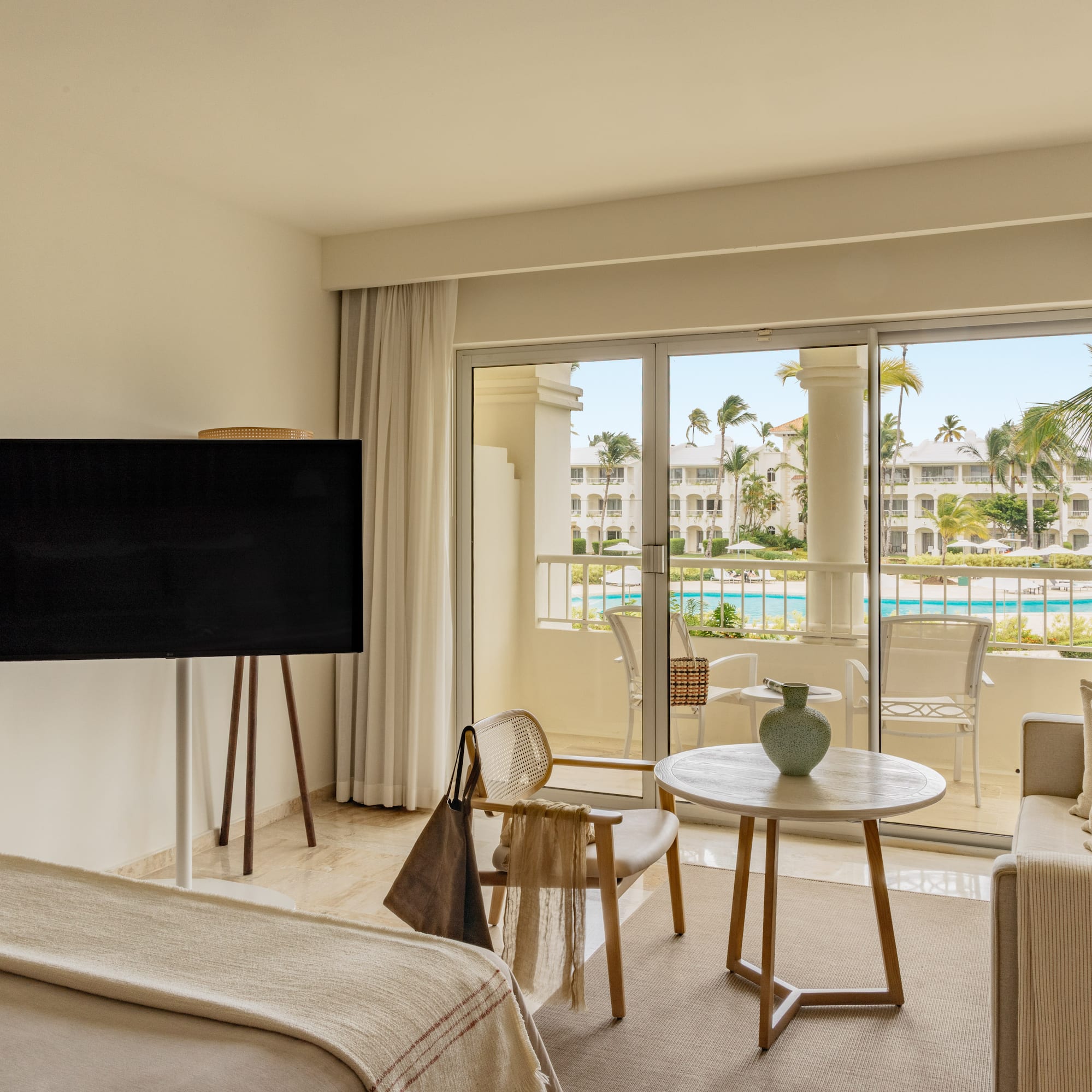 Luxury hotel suite with balcony overlooking resort pool and palm trees.
