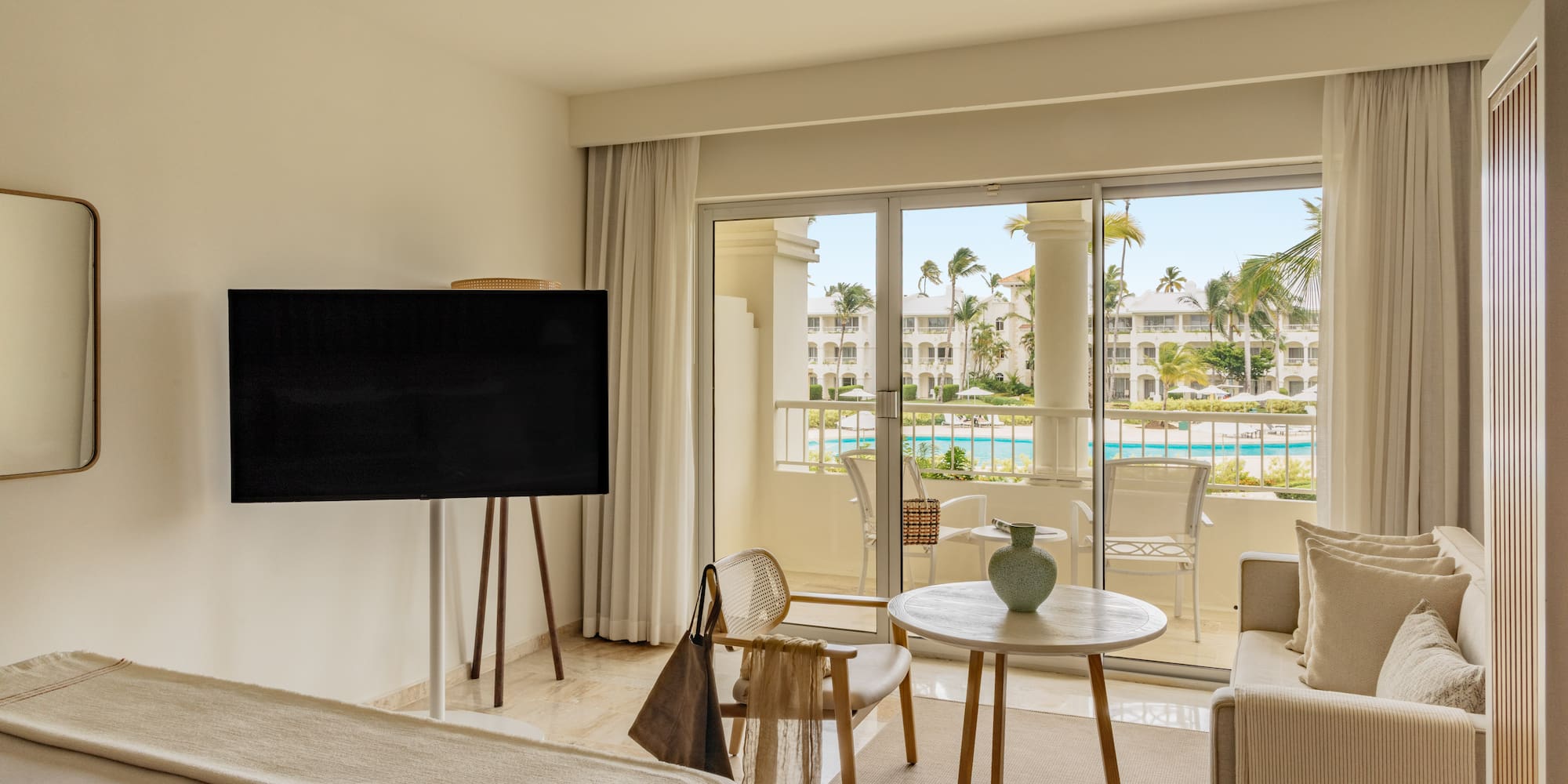 Luxury hotel suite with balcony overlooking resort pool and palm trees.