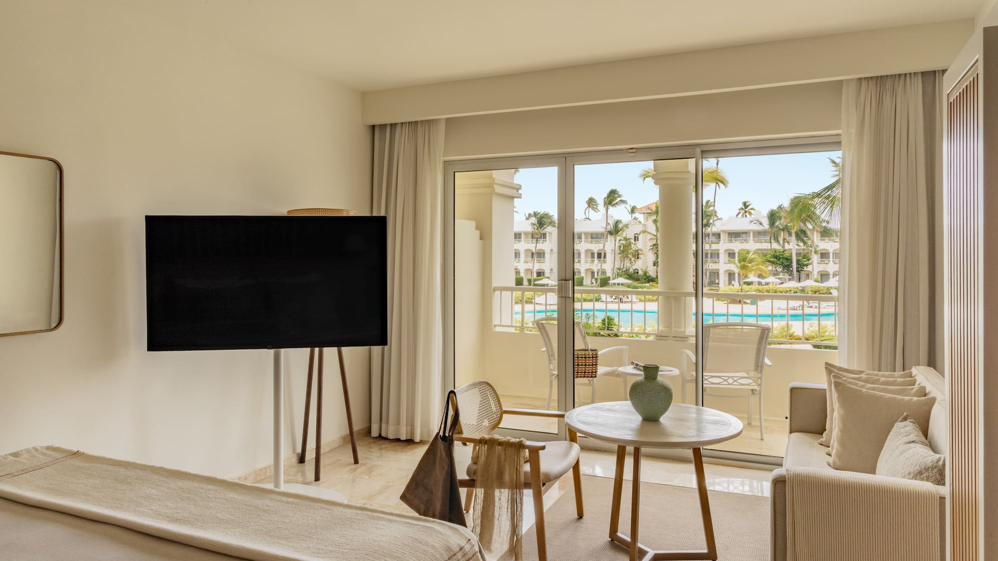 Luxury hotel suite with balcony overlooking resort pool and palm trees.