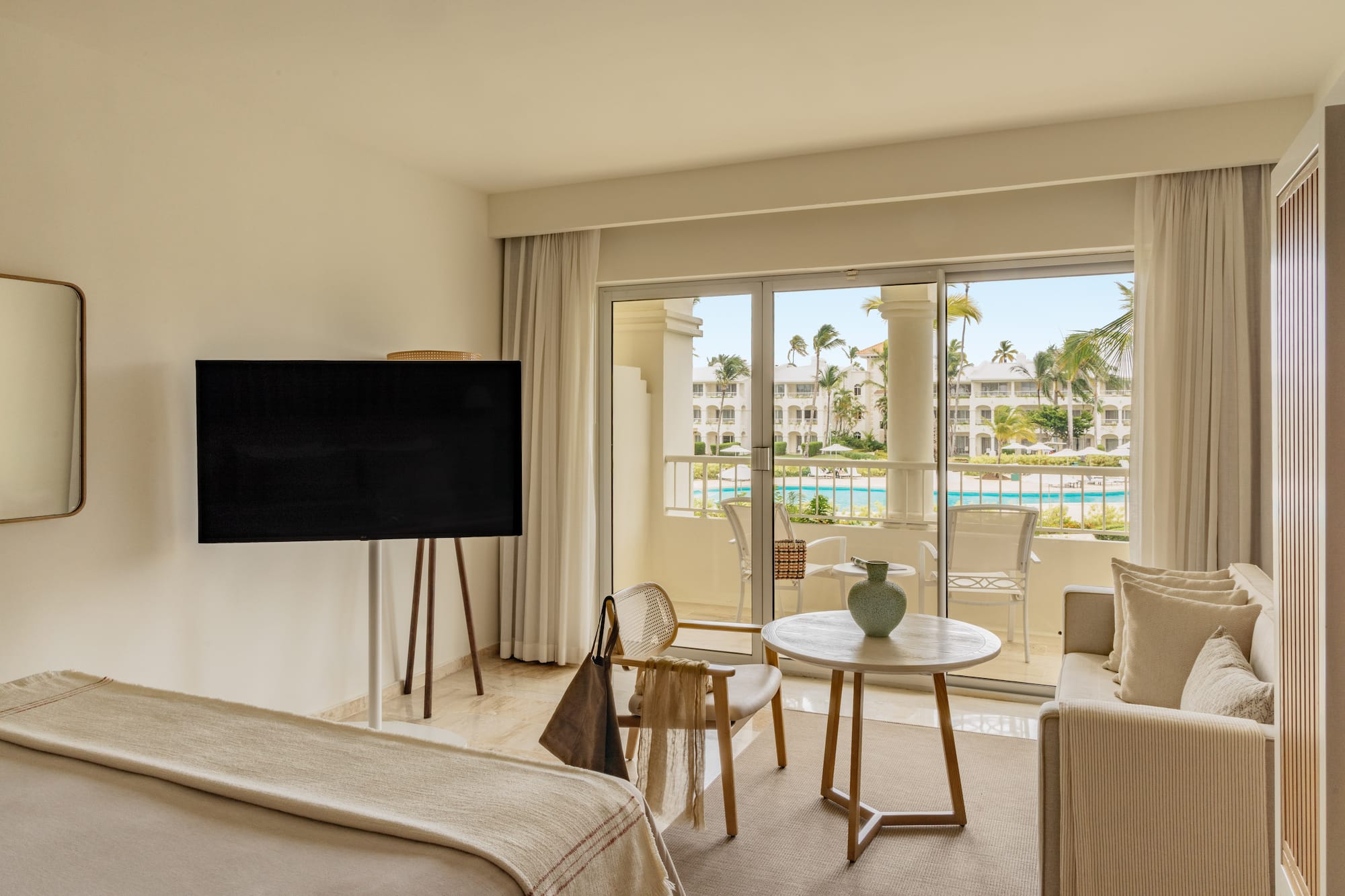 Luxury hotel suite with balcony overlooking resort pool and palm trees.