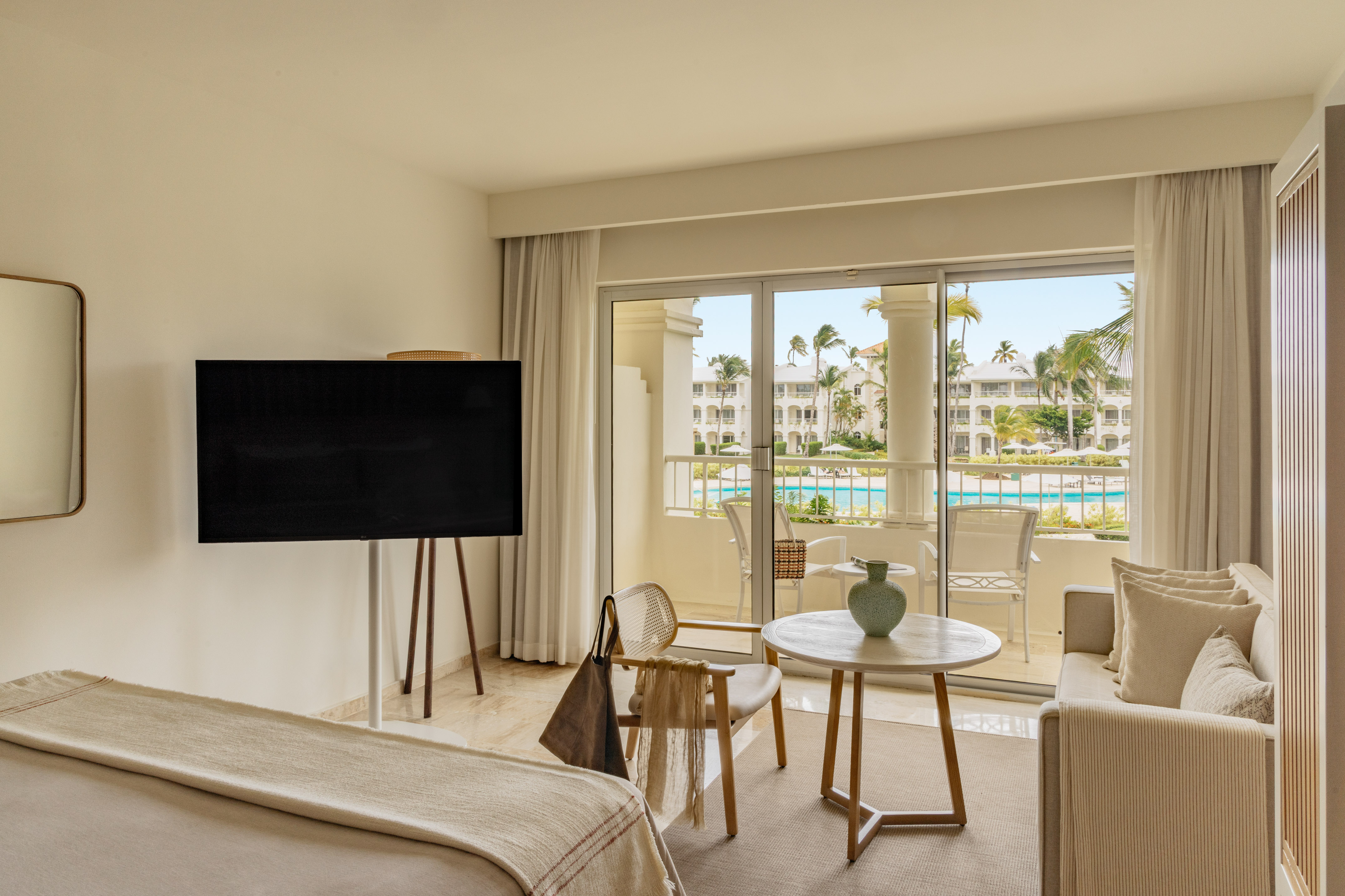 Luxury hotel suite with balcony overlooking resort pool and palm trees.