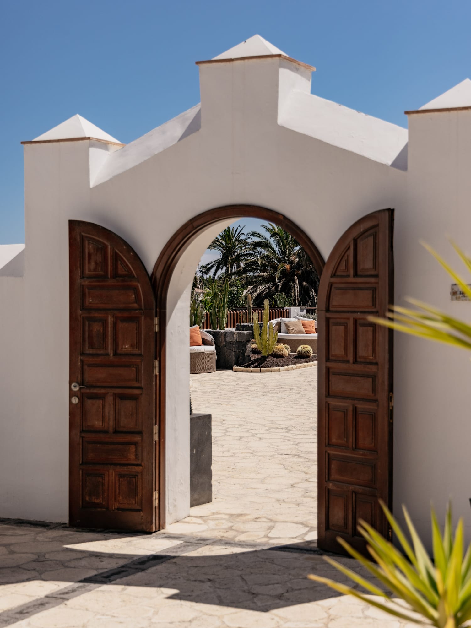 White archway with open wooden doors to a sunny patio with cacti and palm trees