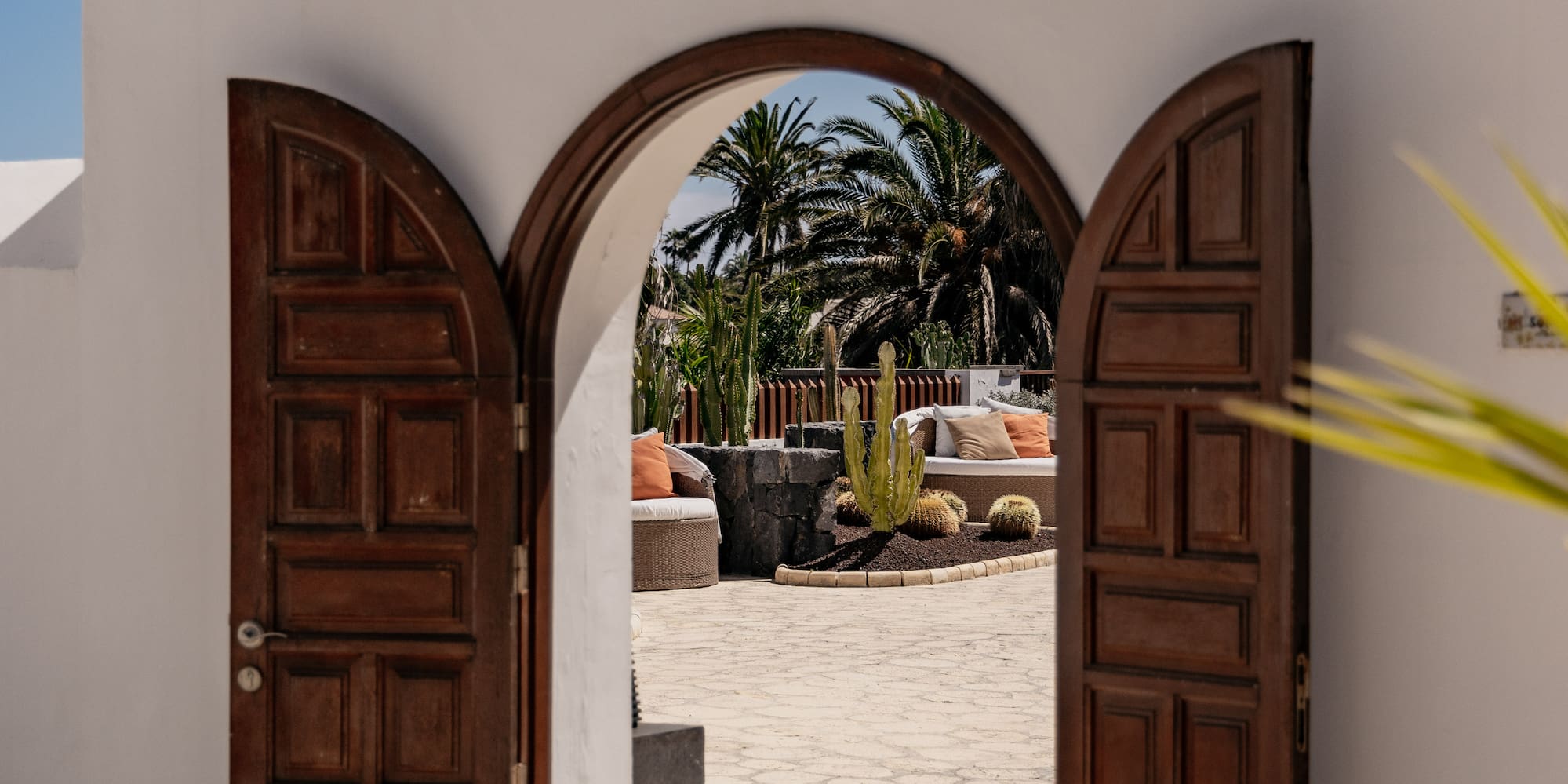 Open arched wooden doors reveal a sunny courtyard with palms, cacti, and outdoor seating.
