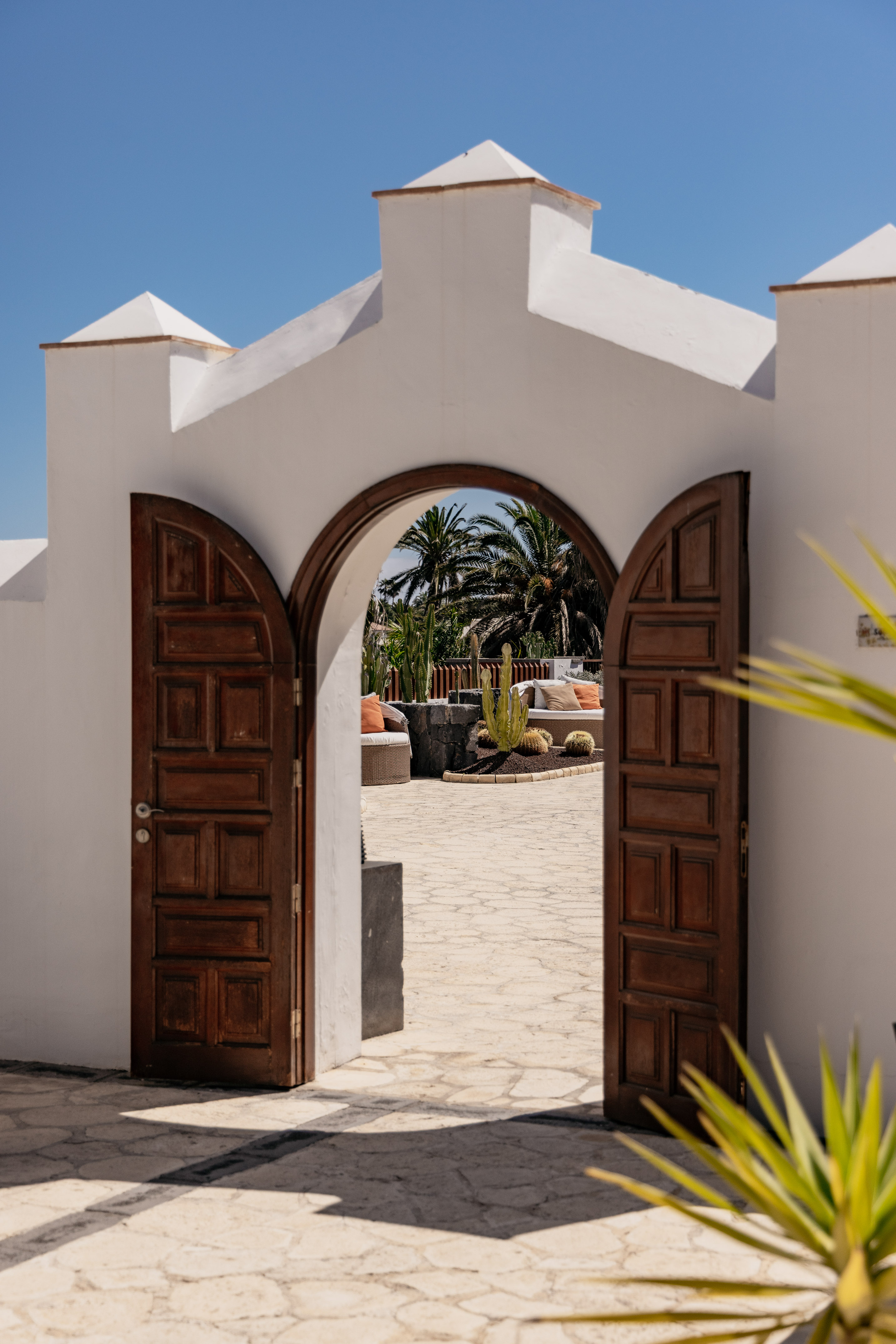 White archway with open wooden doors to a sunny patio with cacti and palm trees