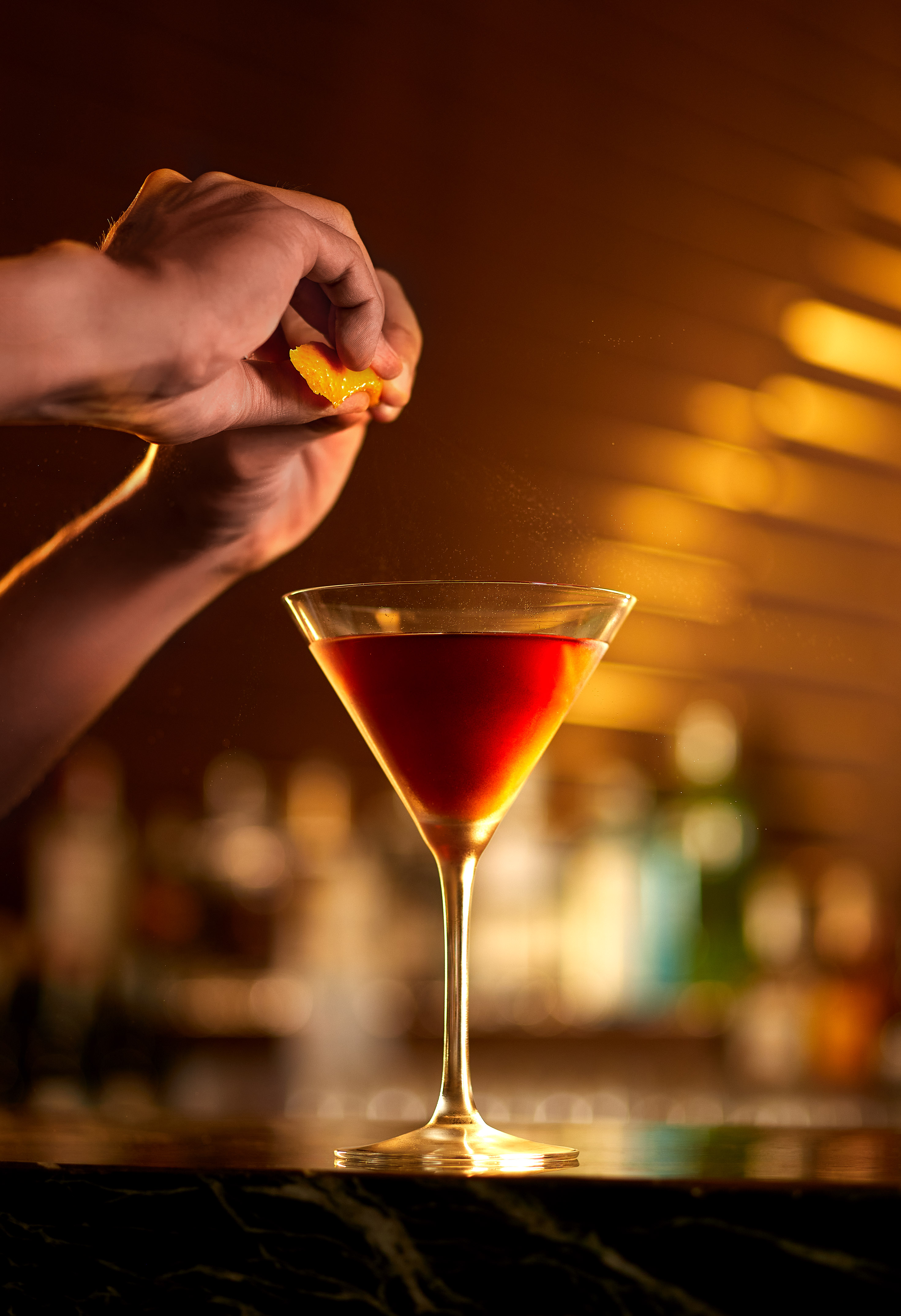 Mixologist garnishing red cocktail with orange peel zest in martini glass.