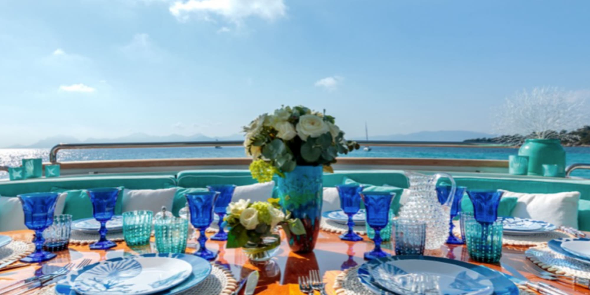 Elegant yacht dining table set with blue dinnerware, flowers, and sea view.