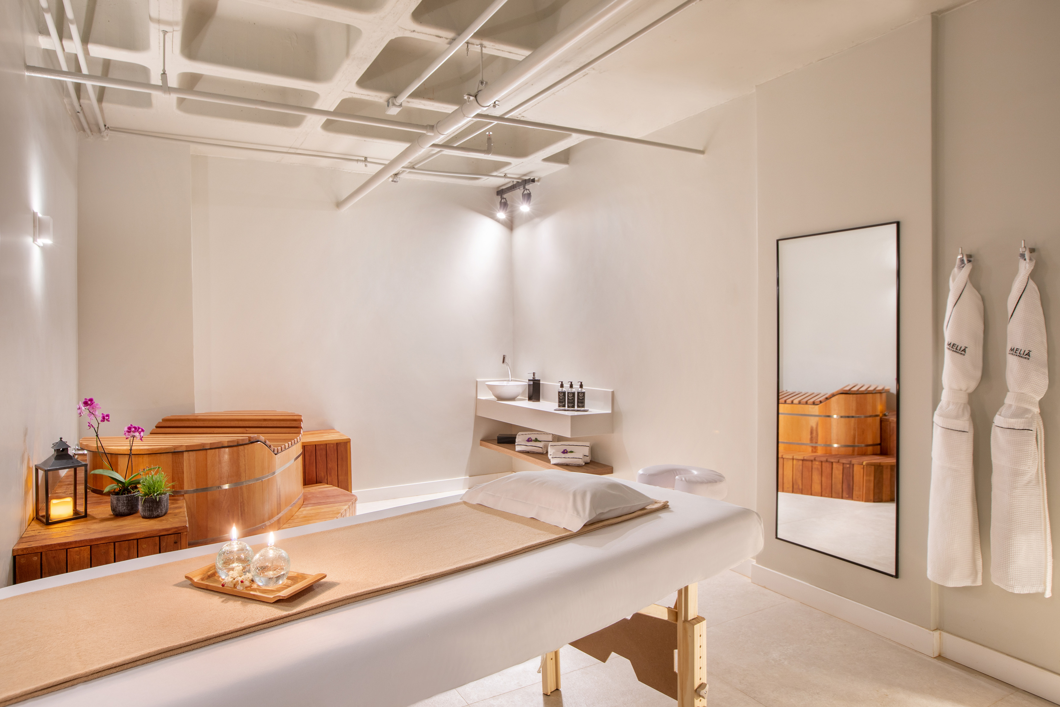 Modern spa treatment room with massage bed, wooden soaking tub, and hanging robes.