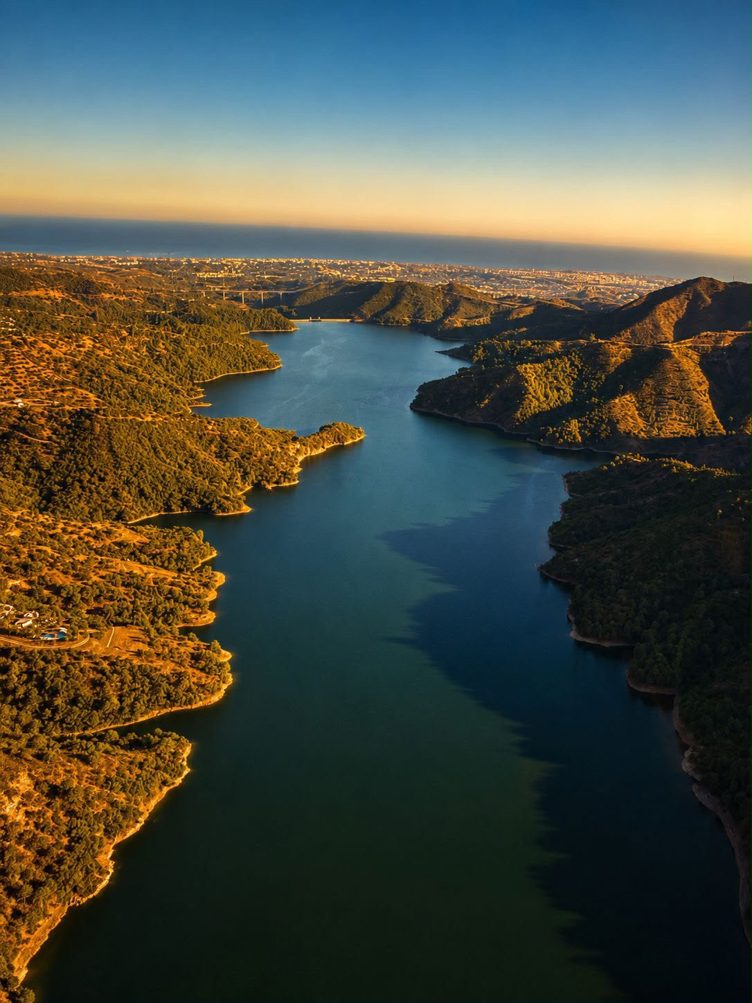Aerial sunset over reservoir, forested mountains, distant city, and ocean.