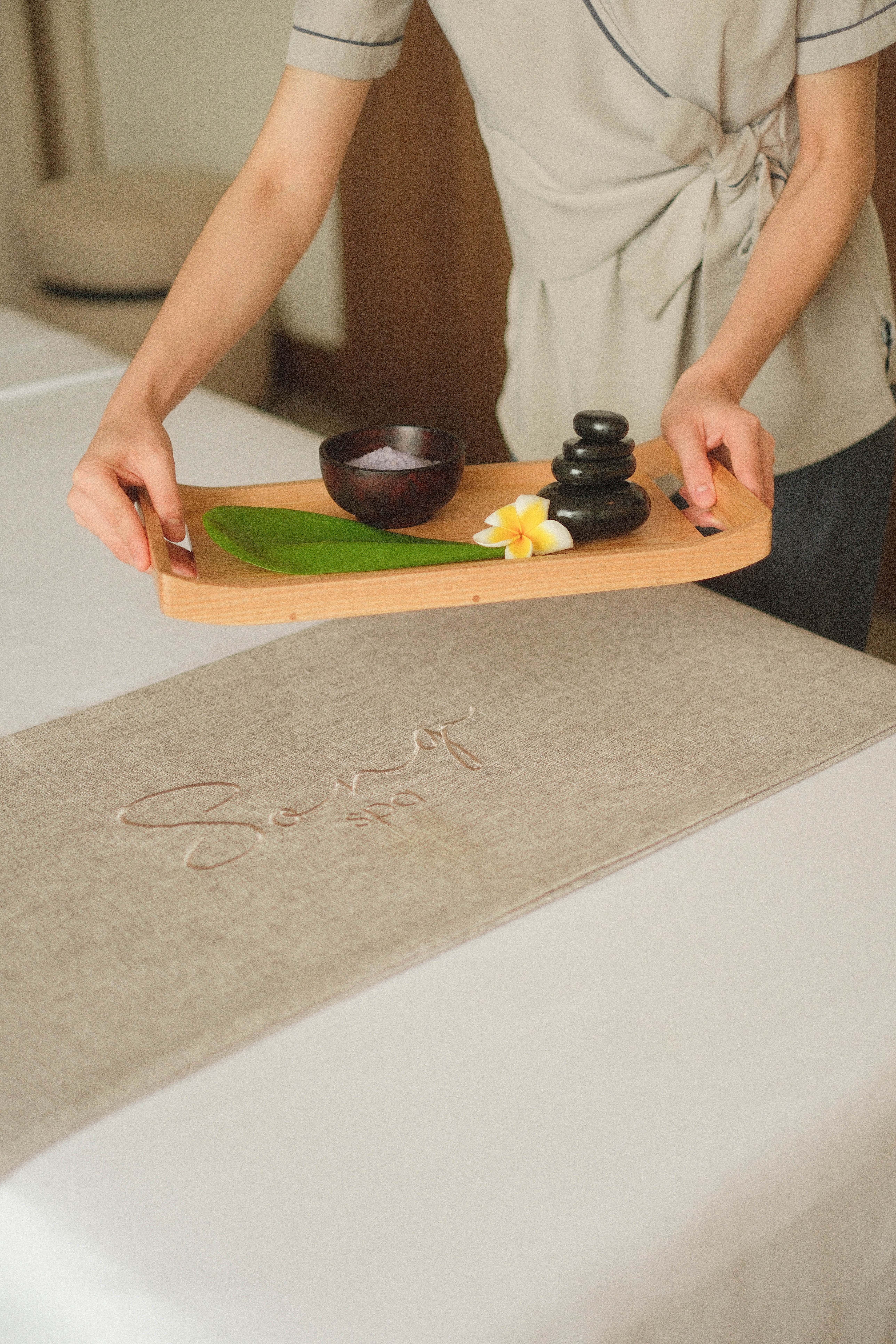 Therapist placing spa tray with hot stones, salts, and plumeria flower.