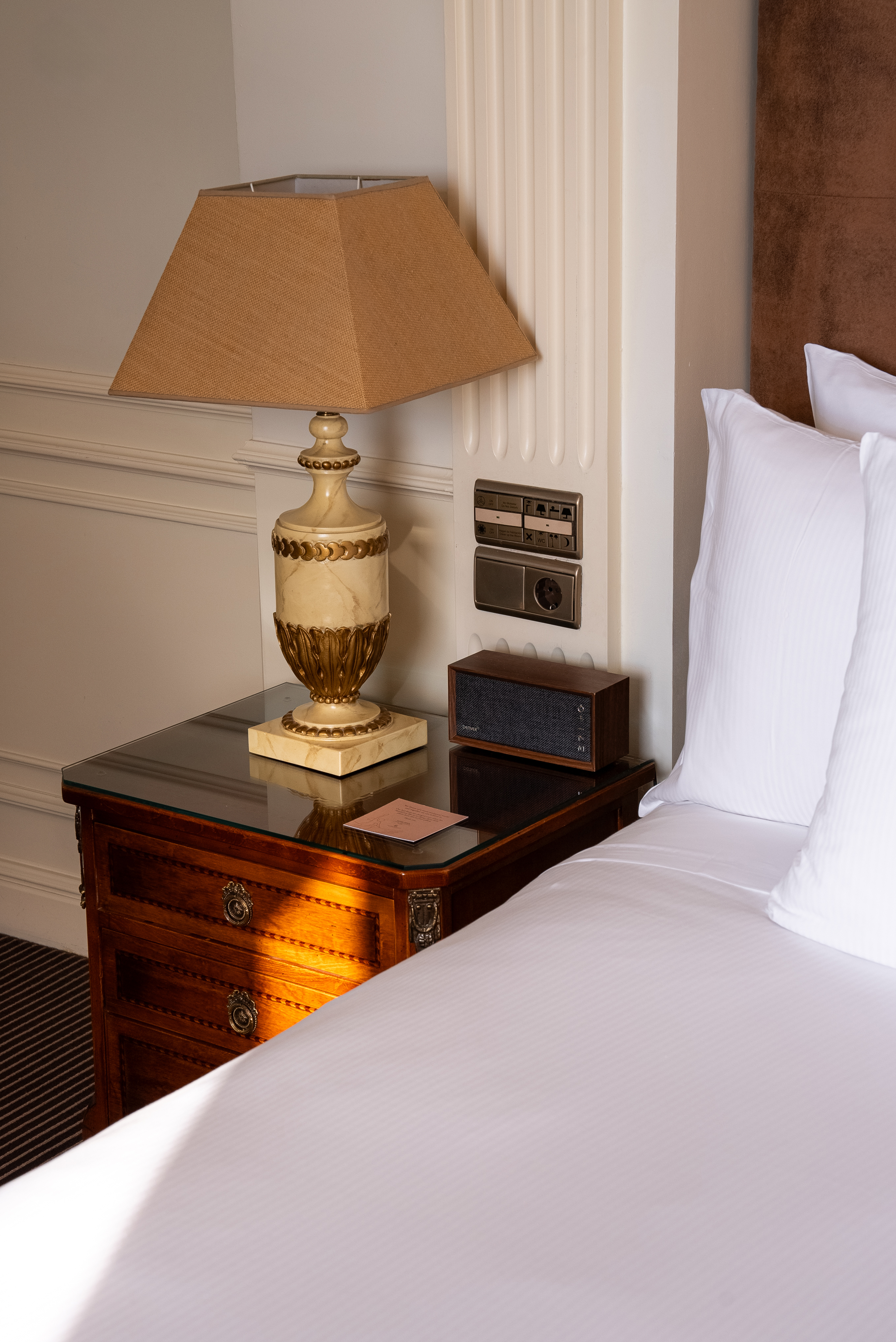 Ornate lamp, wooden nightstand, white hotel bedding, and speaker.