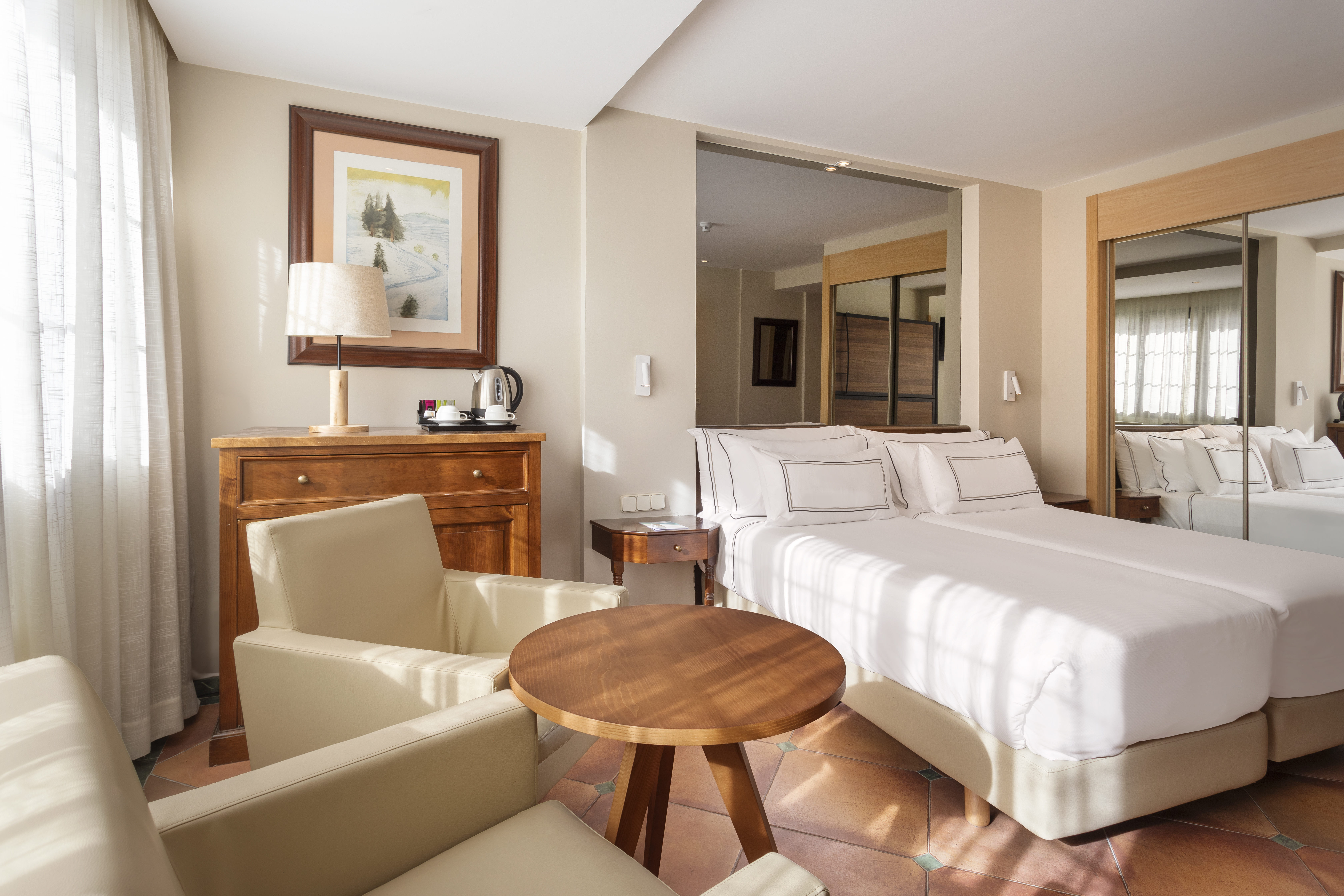 Hotel bedroom with twin beds, two armchairs, wooden table, dresser, and mirrors.