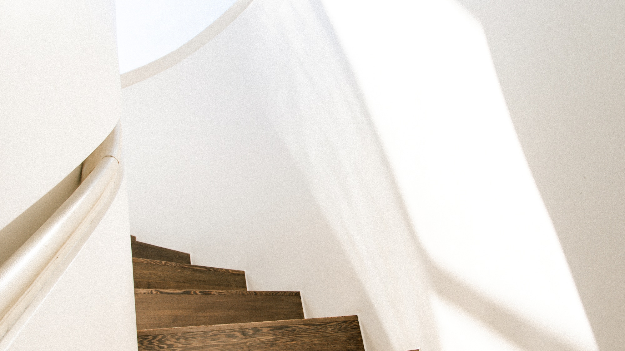 Sunlit dark wood spiral staircase, white walls, modern interior design.