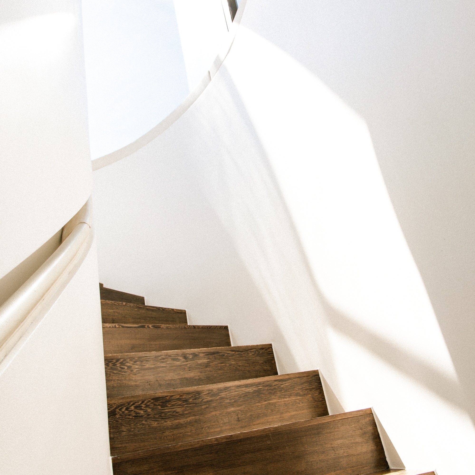 Sunlit dark wood spiral staircase, white walls, modern interior design.