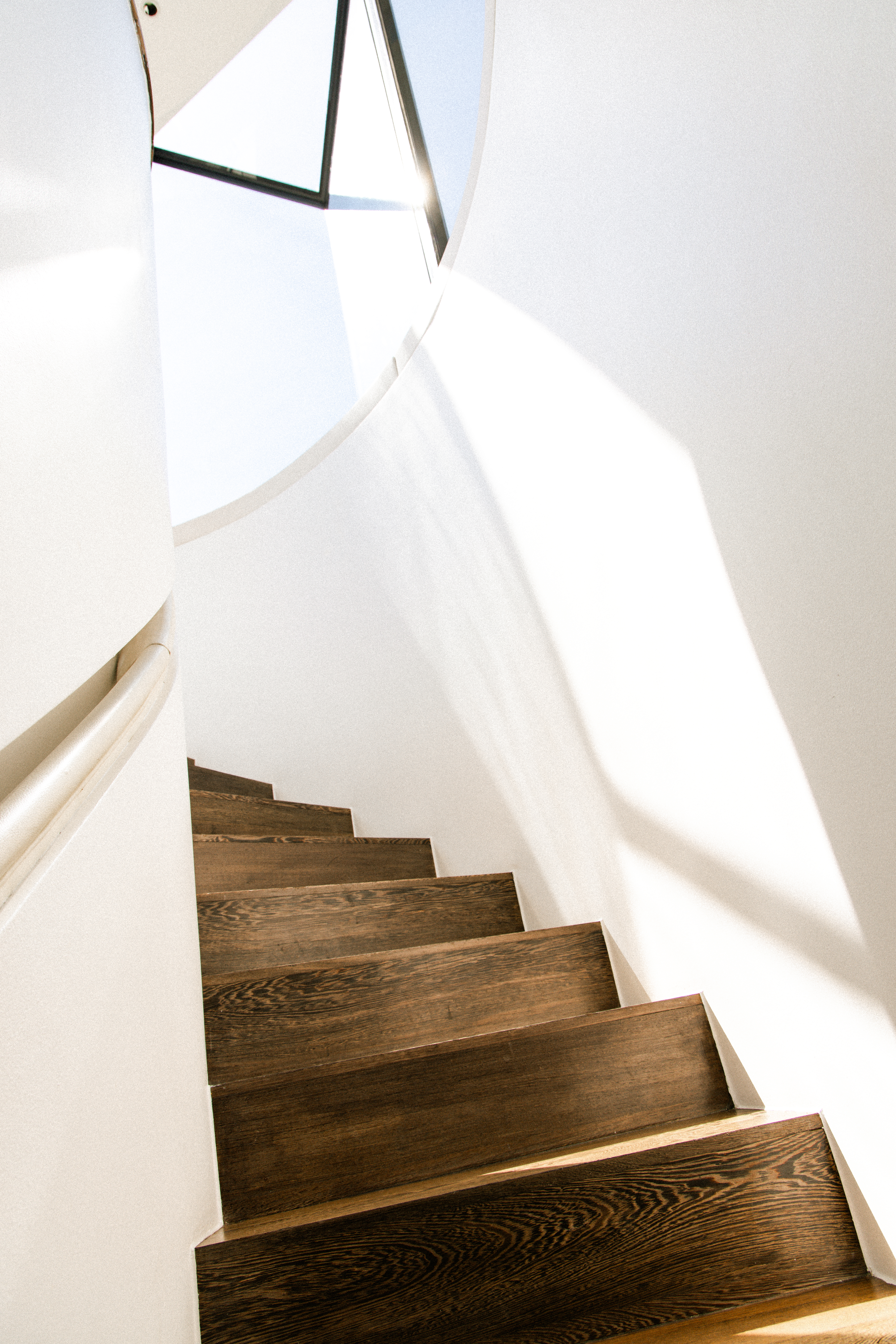 Sunlit dark wood spiral staircase, white walls, modern interior design.
