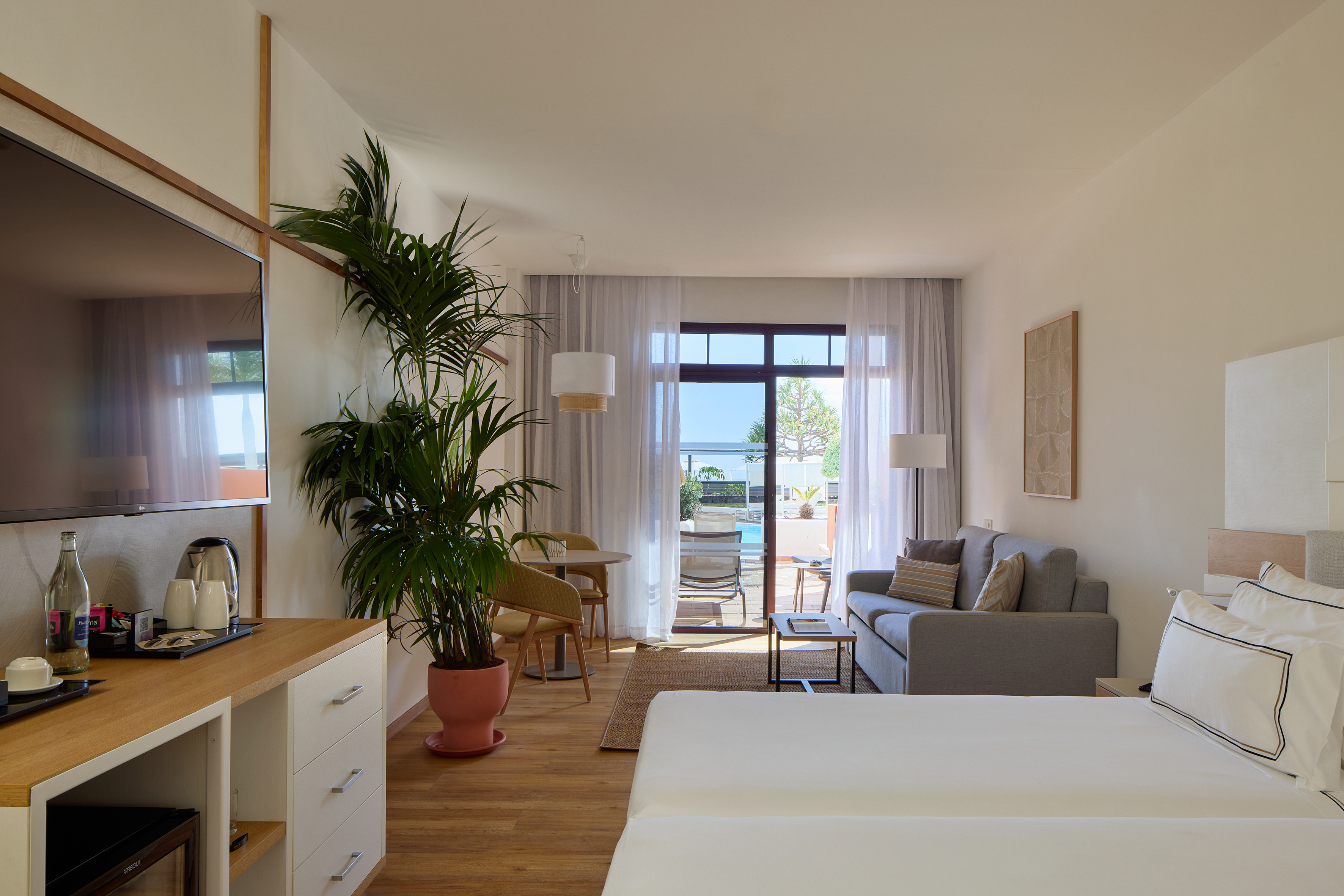 Bright hotel suite with bedroom, living area, balcony, pool, and sea view.