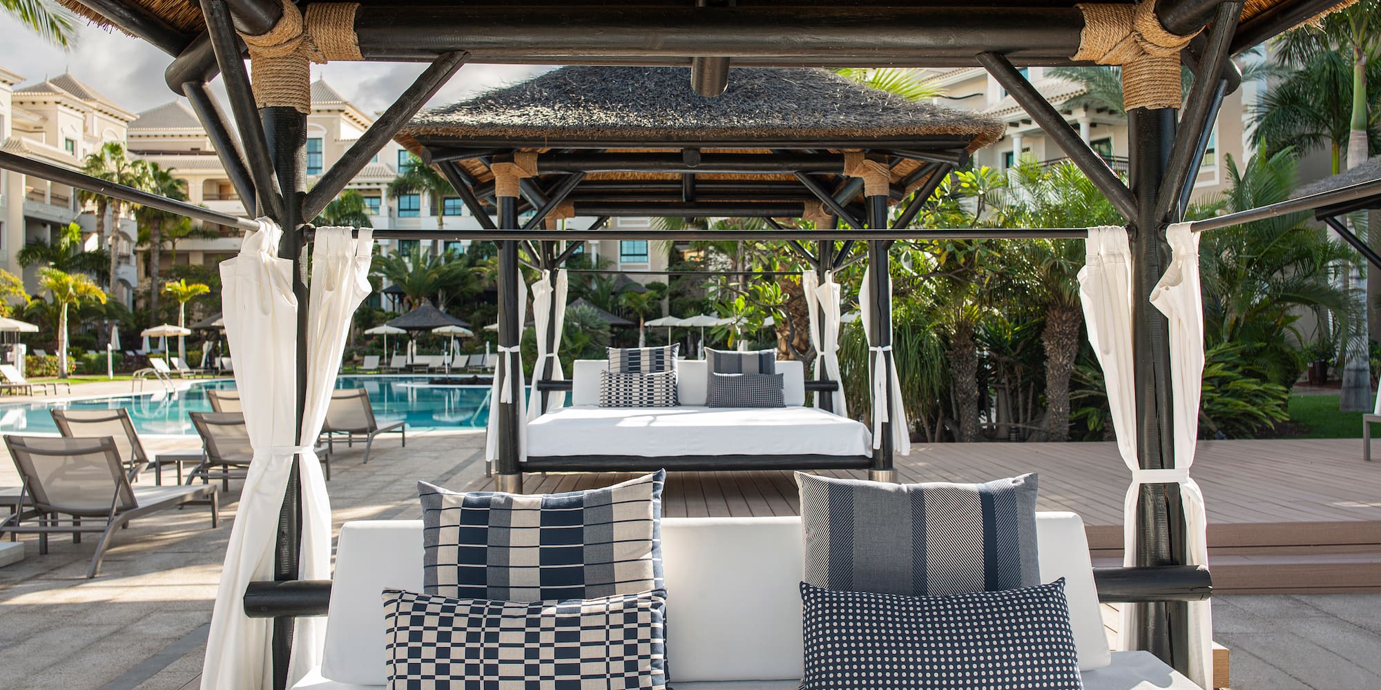 Luxury resort cabana with thatched roof, daybeds, and swimming pool.