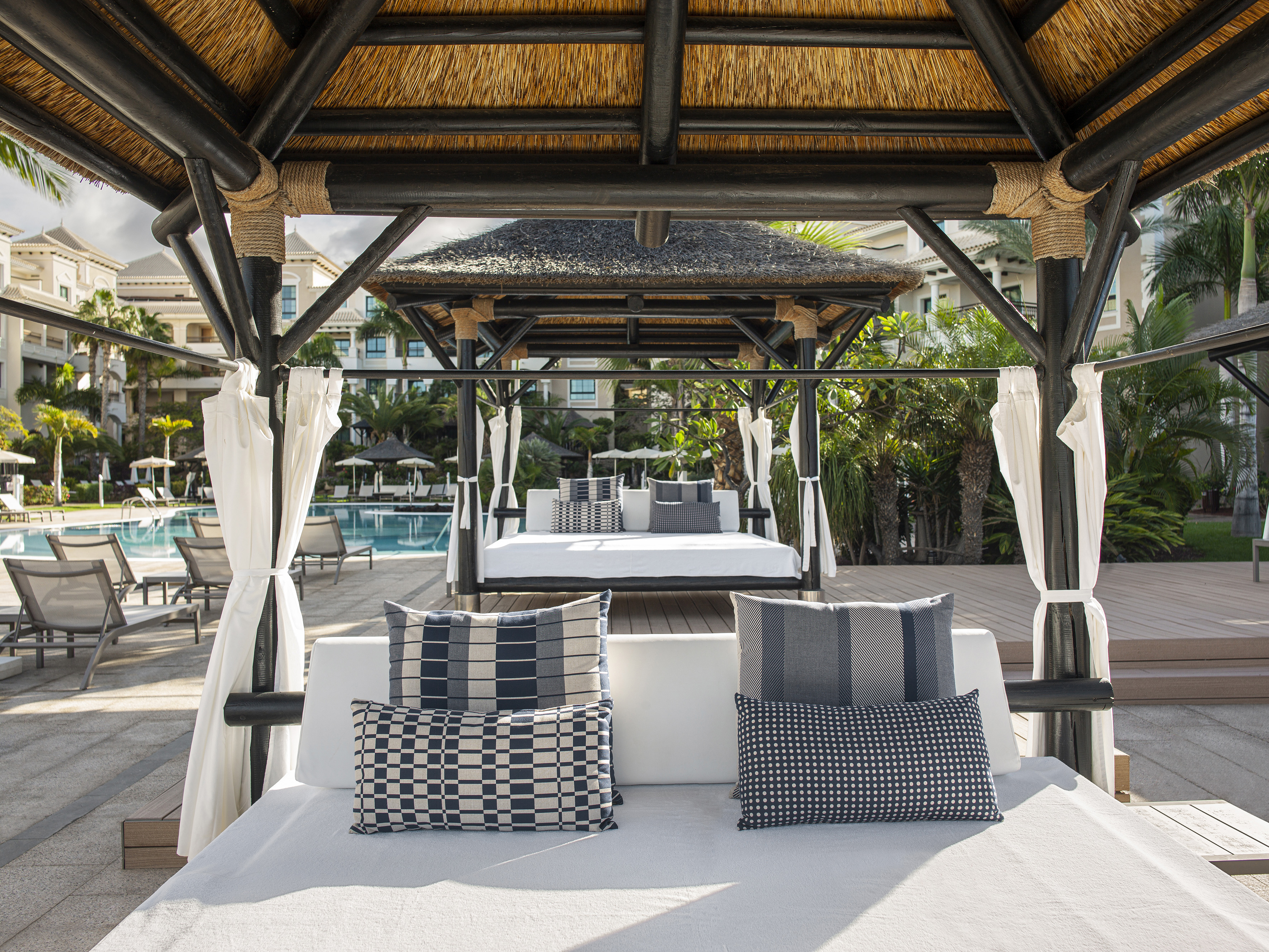 Luxury resort cabana with thatched roof, daybeds, and swimming pool.