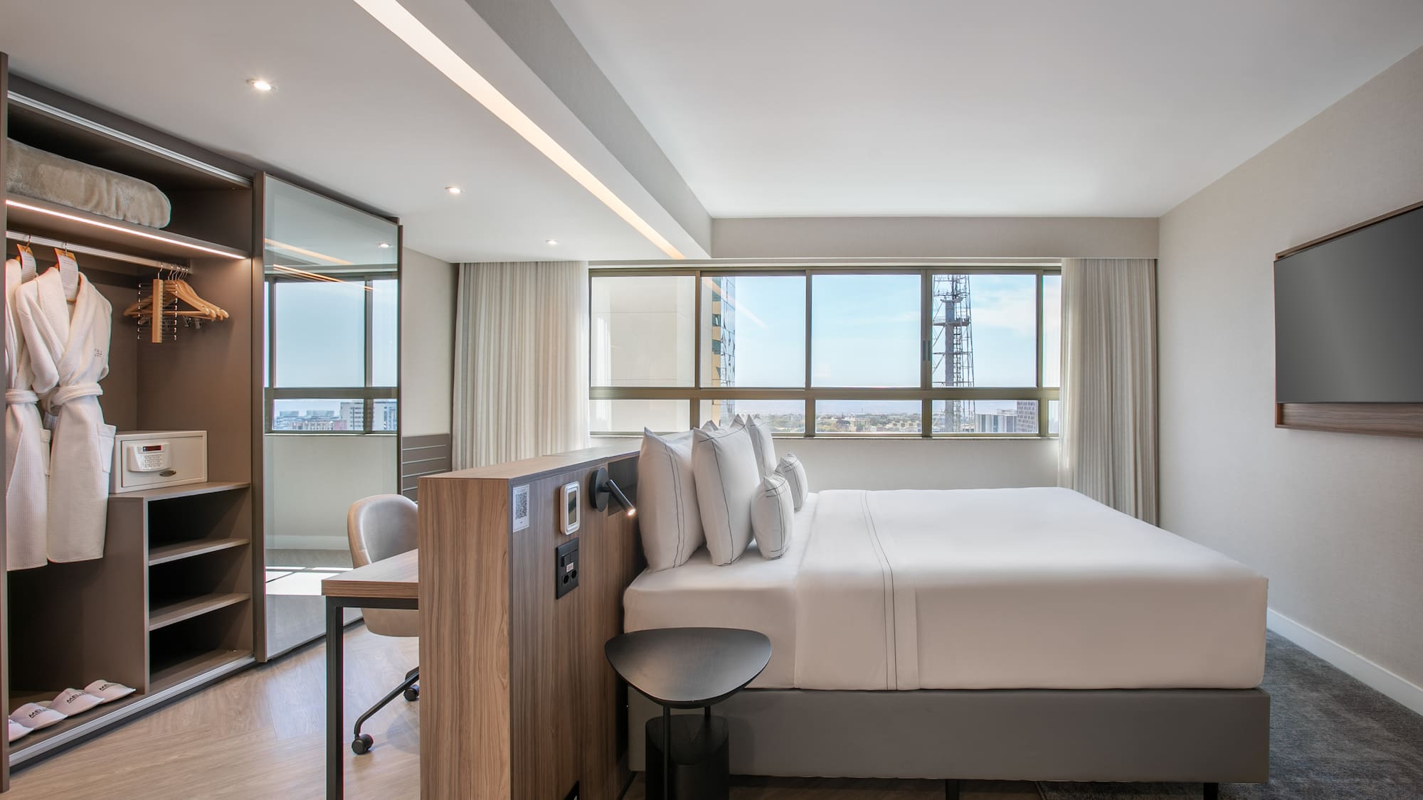 Modern hotel room with comfortable bed, city view window, and desk workspace.