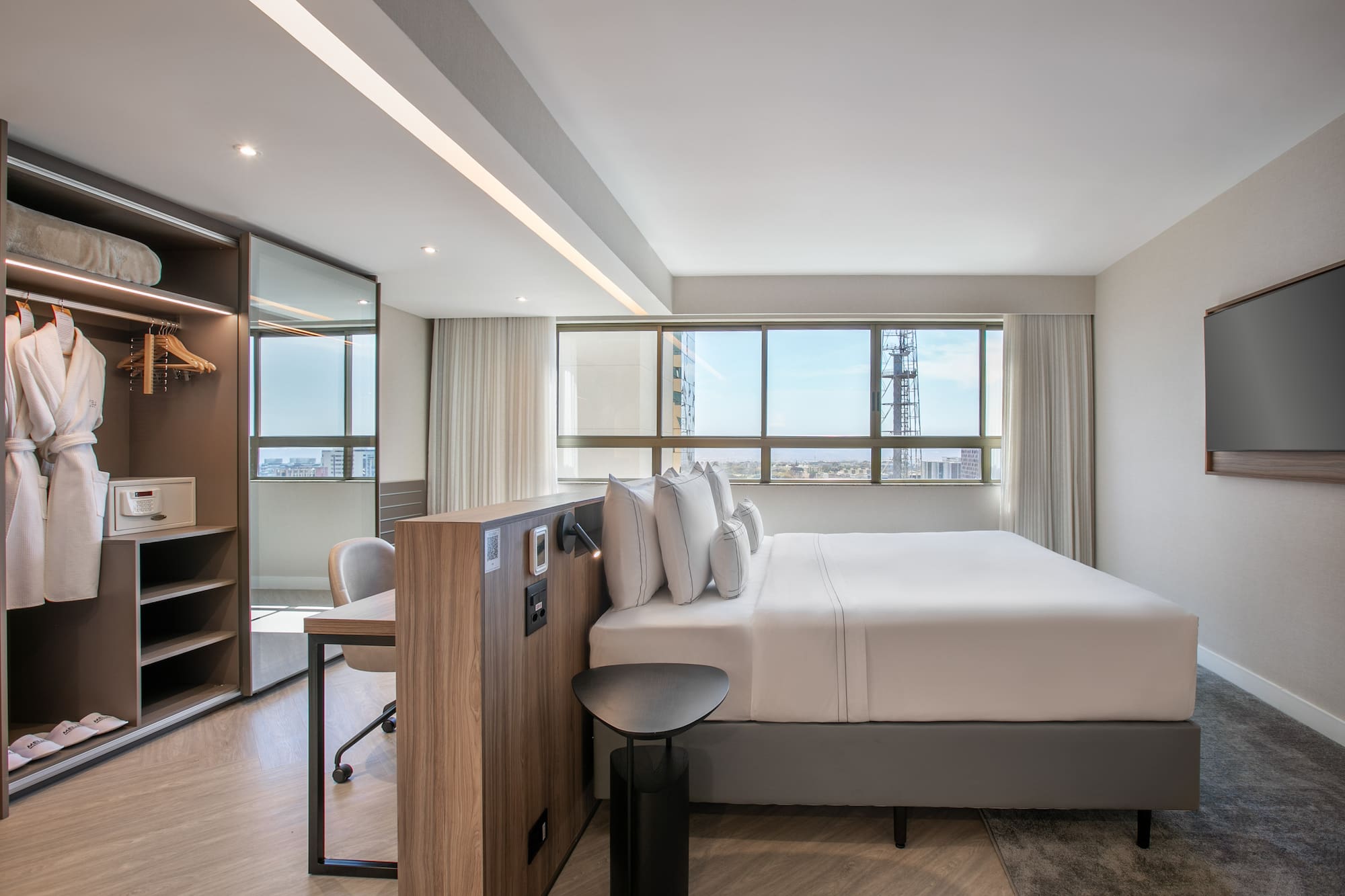 Modern hotel bedroom with city view, king bed, desk, and open closet.