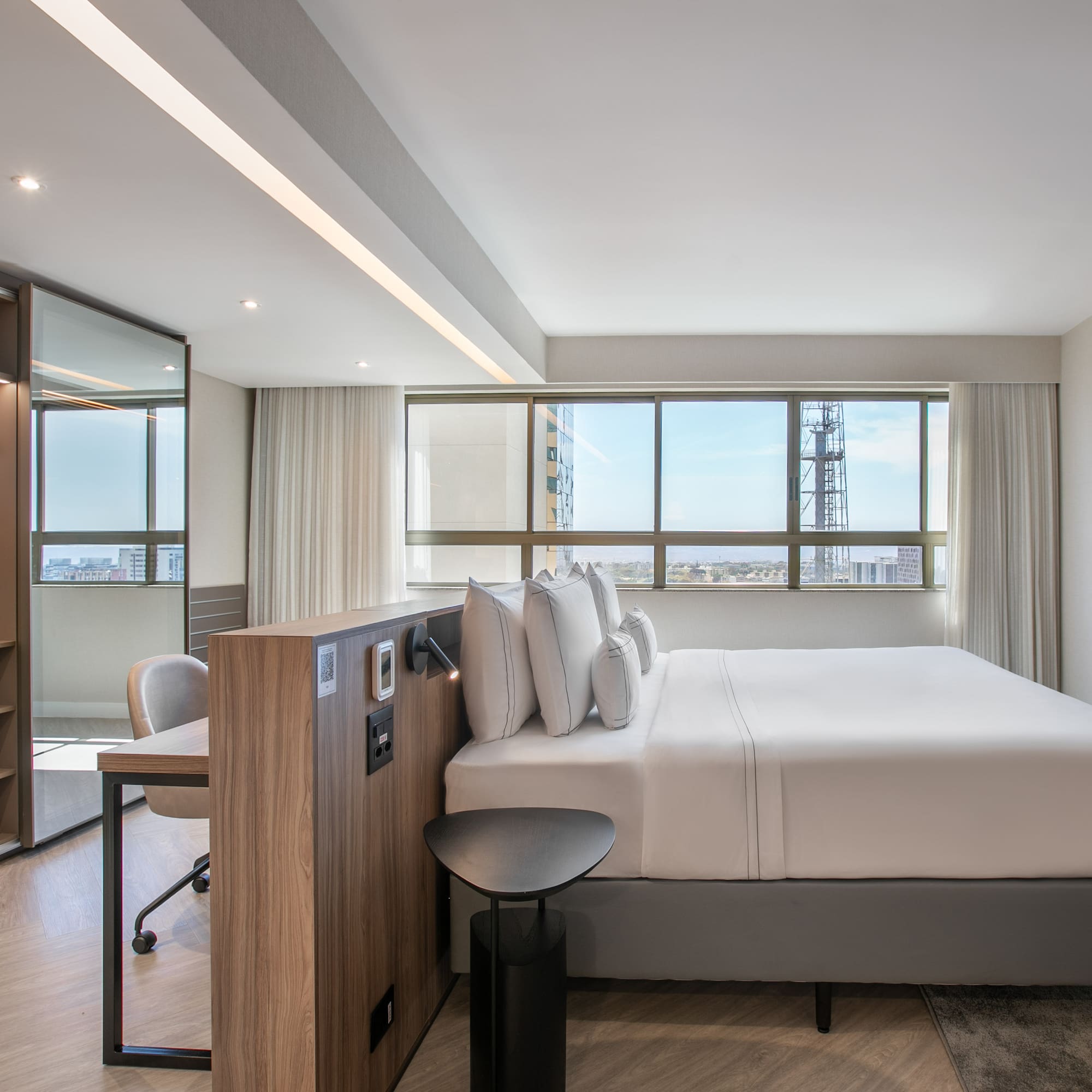 Modern hotel bedroom with city view, king bed, desk, and open closet.