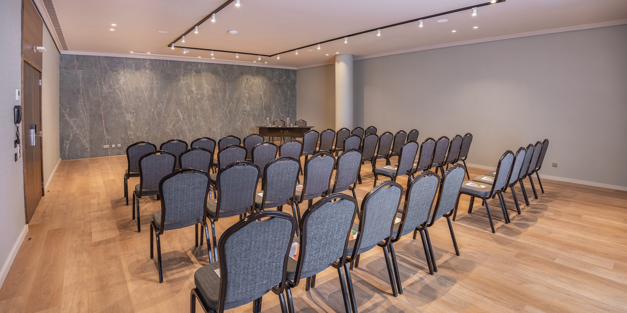 Hotel meeting room with grey chairs, wooden floor, and marble accent wall.