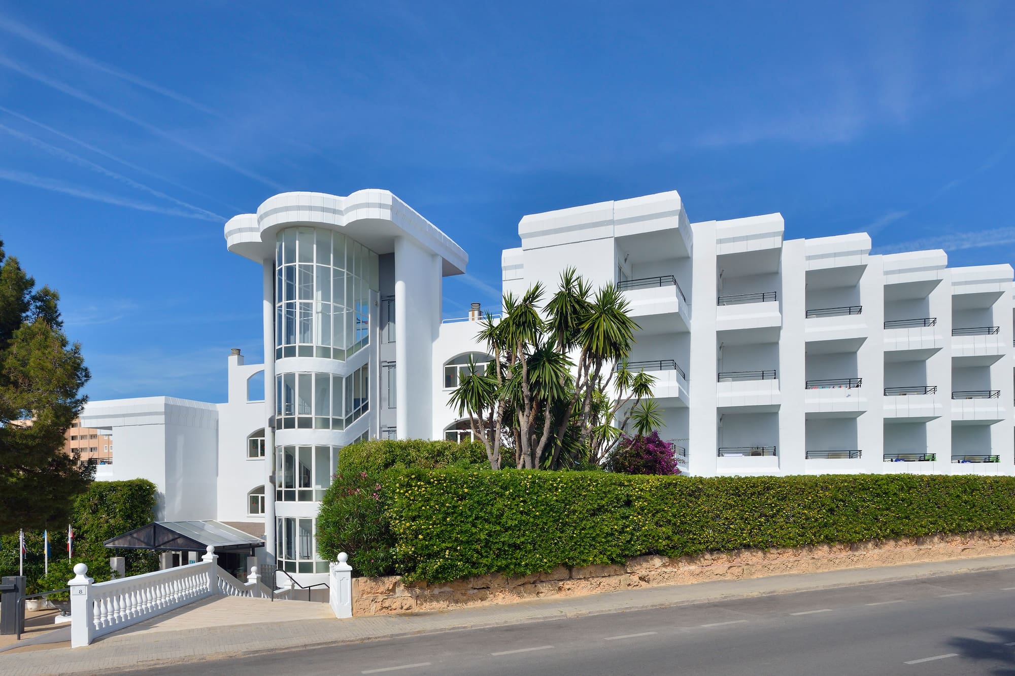 White hotel building with curved glass tower, balconies, palm trees, and hedges.