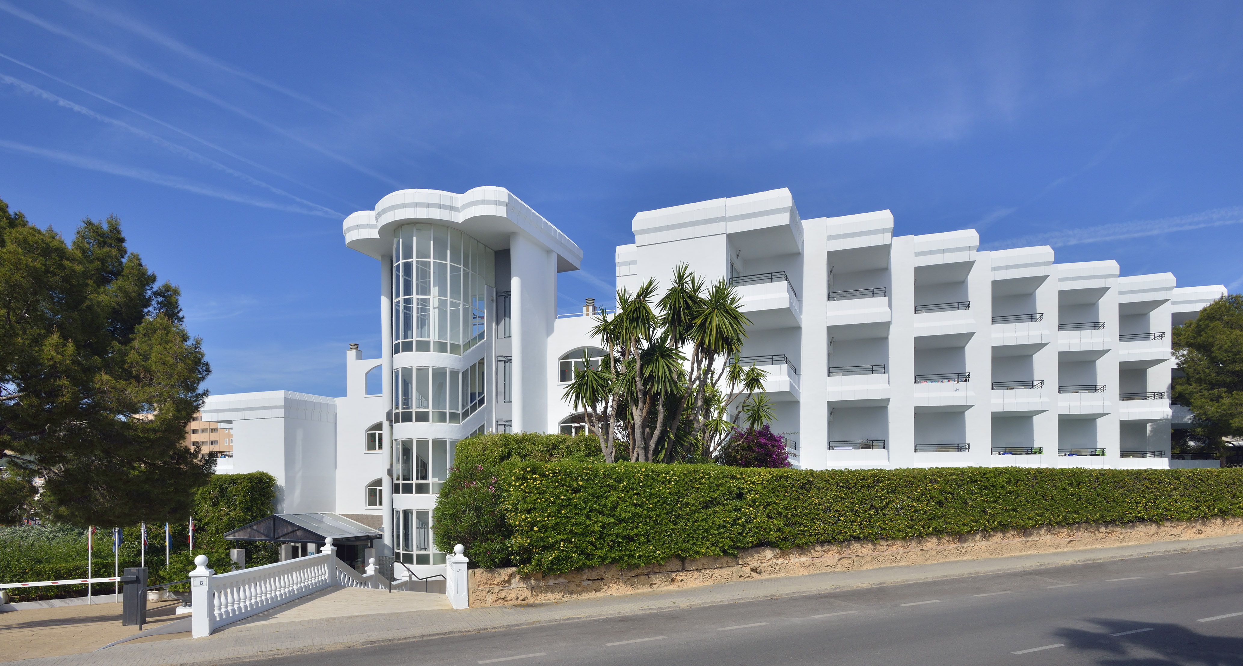 White hotel building with curved glass tower, balconies, palm trees, and hedges.