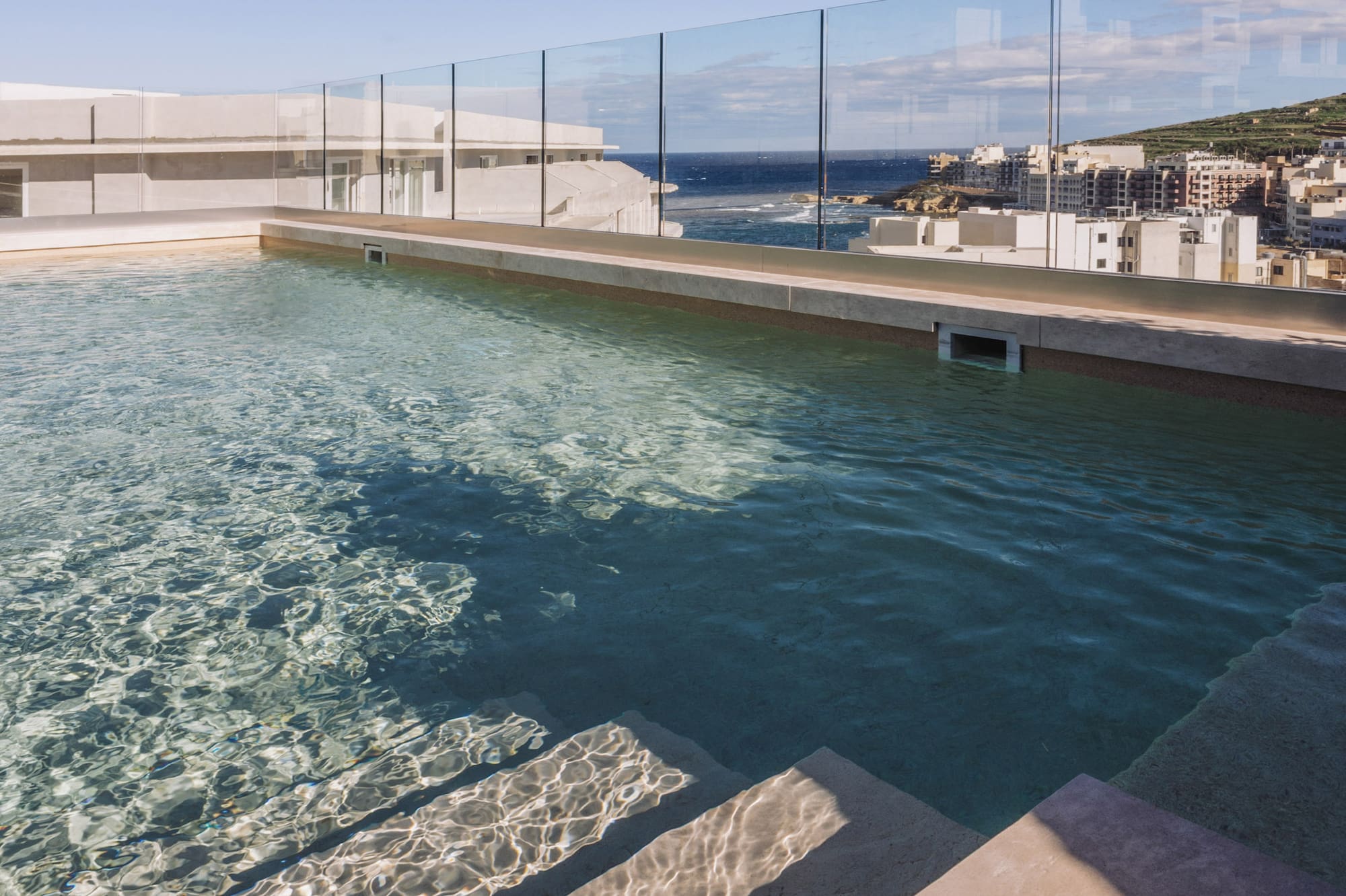 Rooftop pool overlooking ocean and coastal town, with clear water and steps.