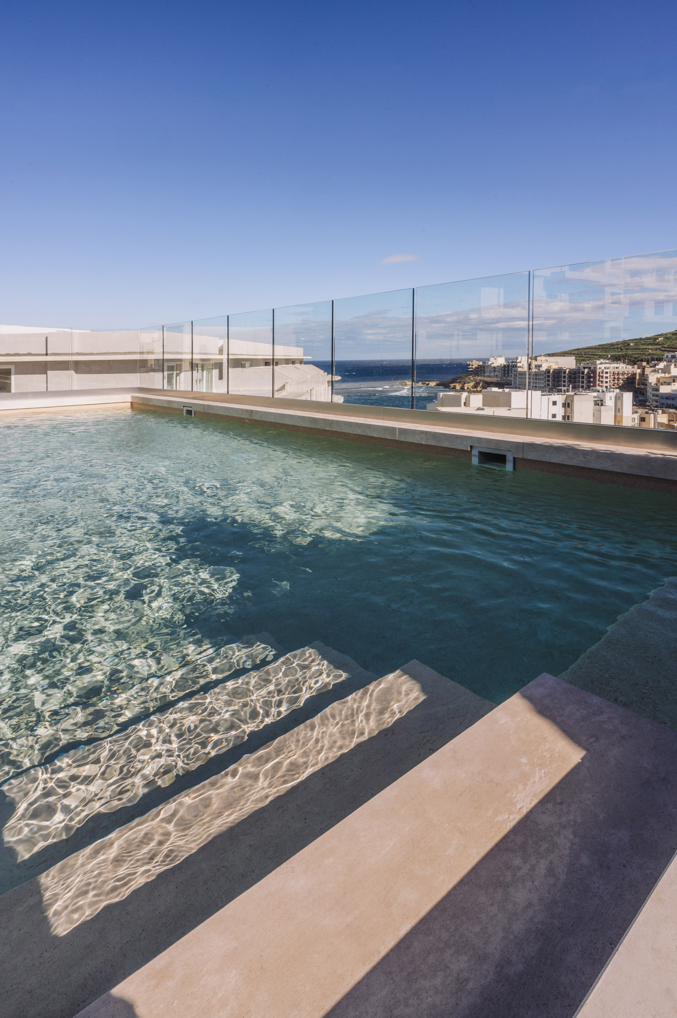 Rooftop pool overlooking ocean and coastal town, with clear water and steps.