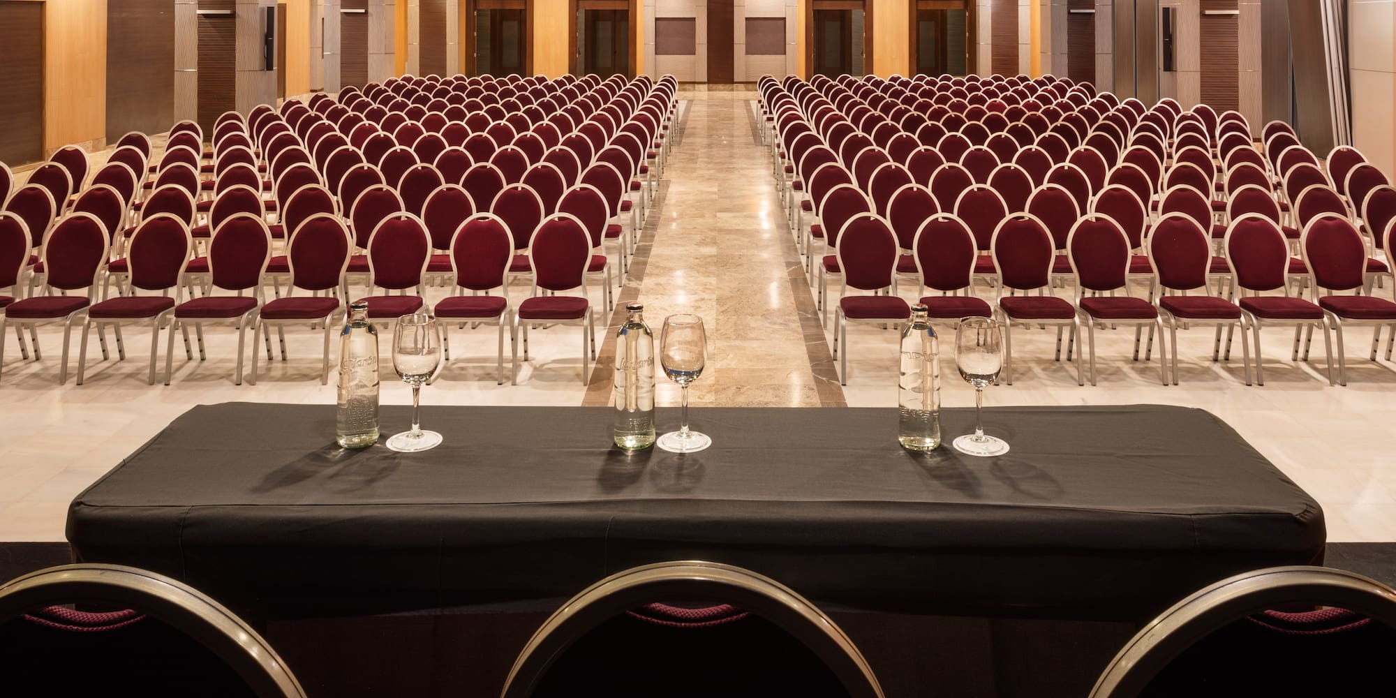 Hotel conference room with rows of red chairs, speaker table with water bottles.