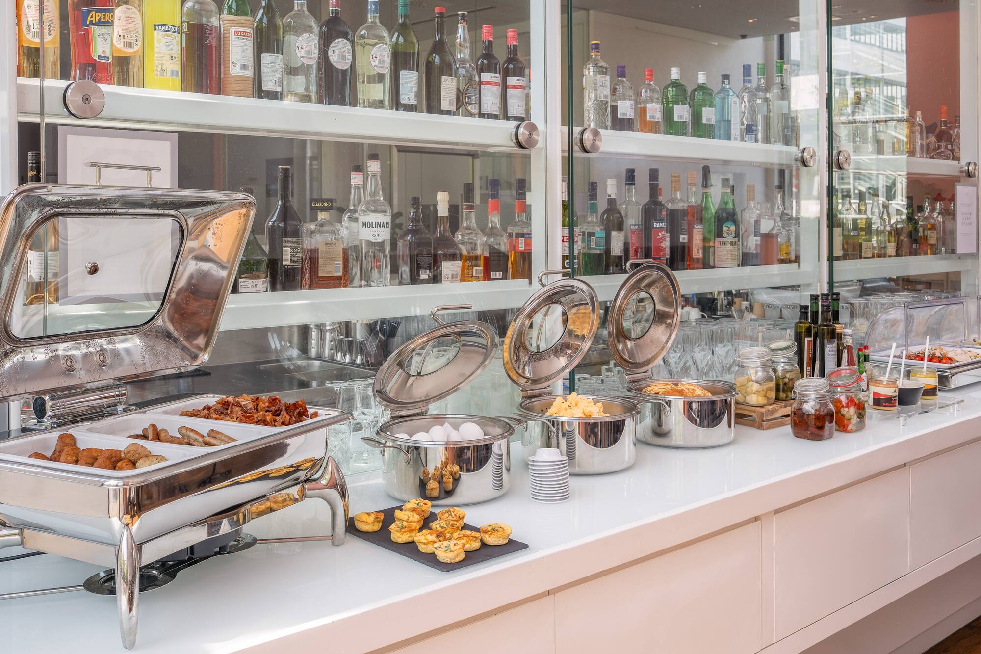 Hotel breakfast buffet with hot food, eggs, quiches, and bar bottles.