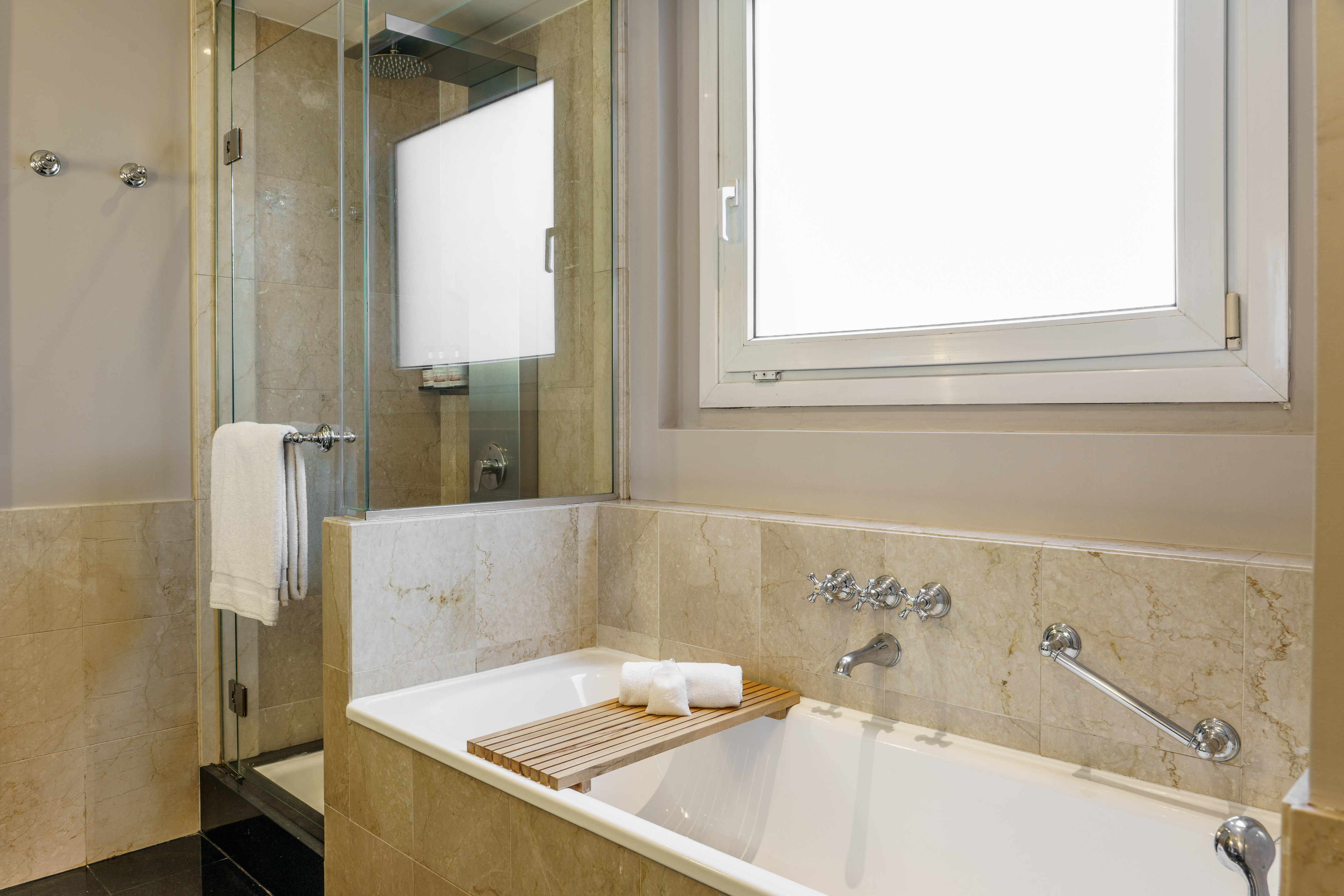 Modern hotel bathroom with marble tub, glass shower, and window.