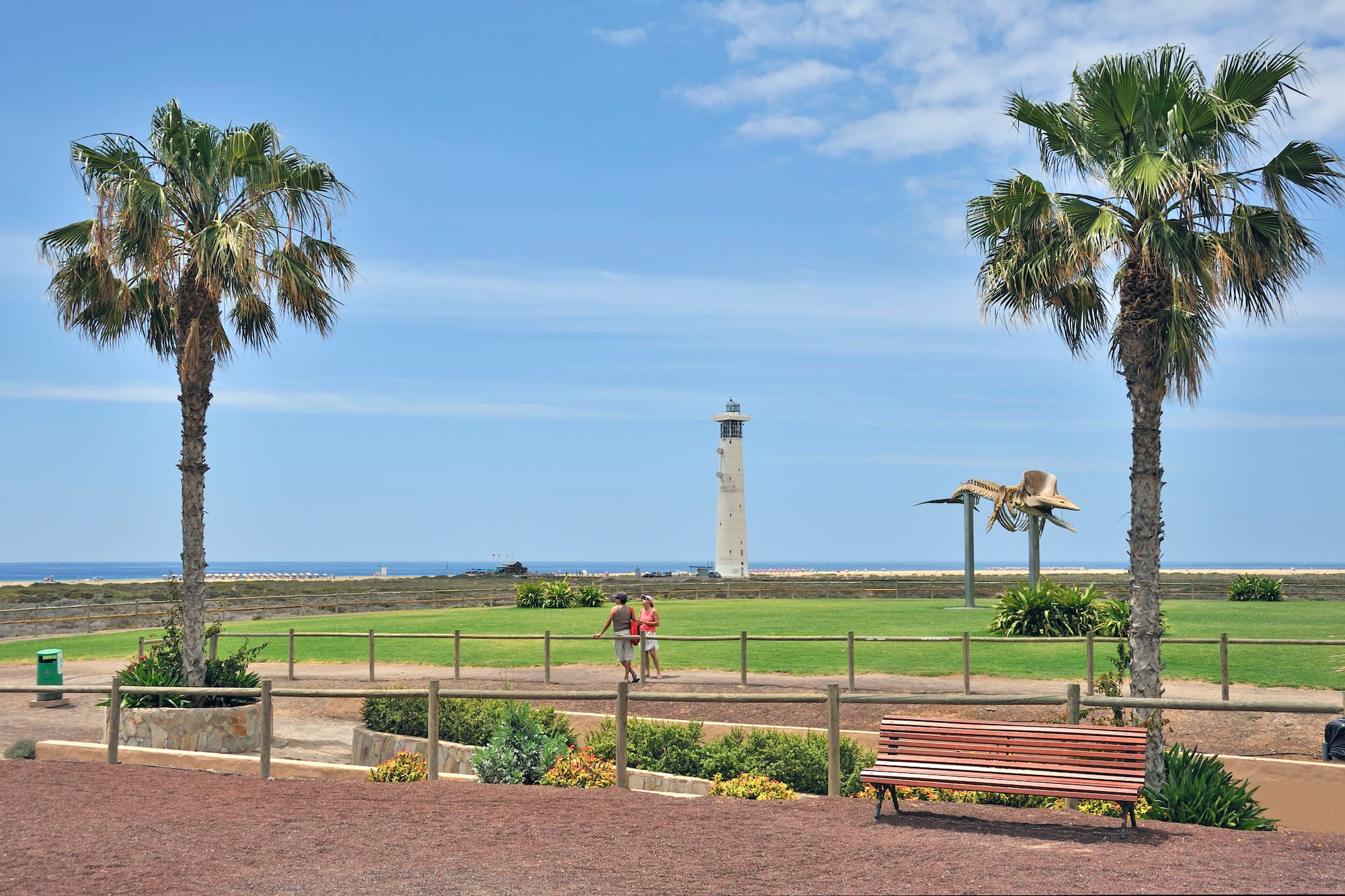 Faro, palmeras y esqueleto de ballena, con playa, césped y personas.