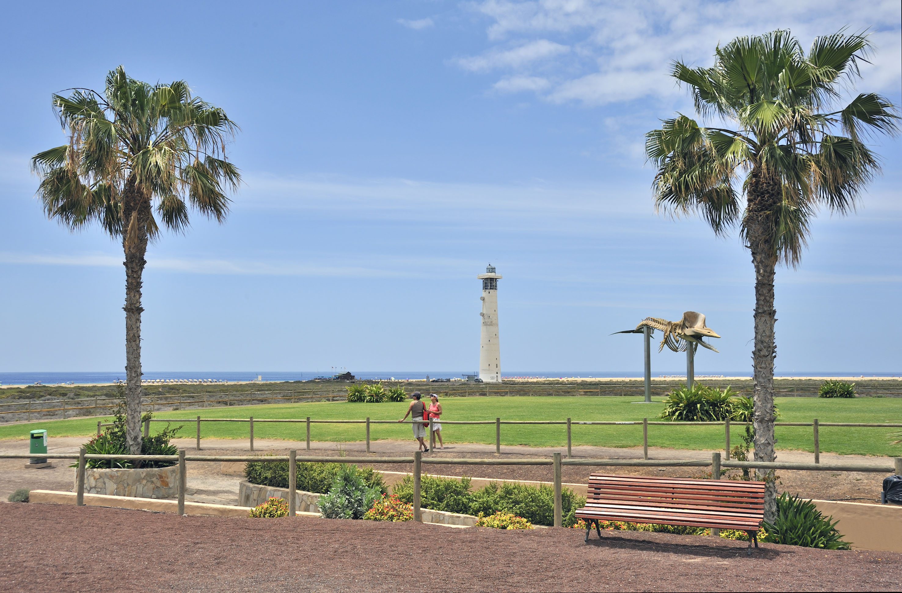 Faro, palmeras y esqueleto de ballena, con playa, césped y personas.