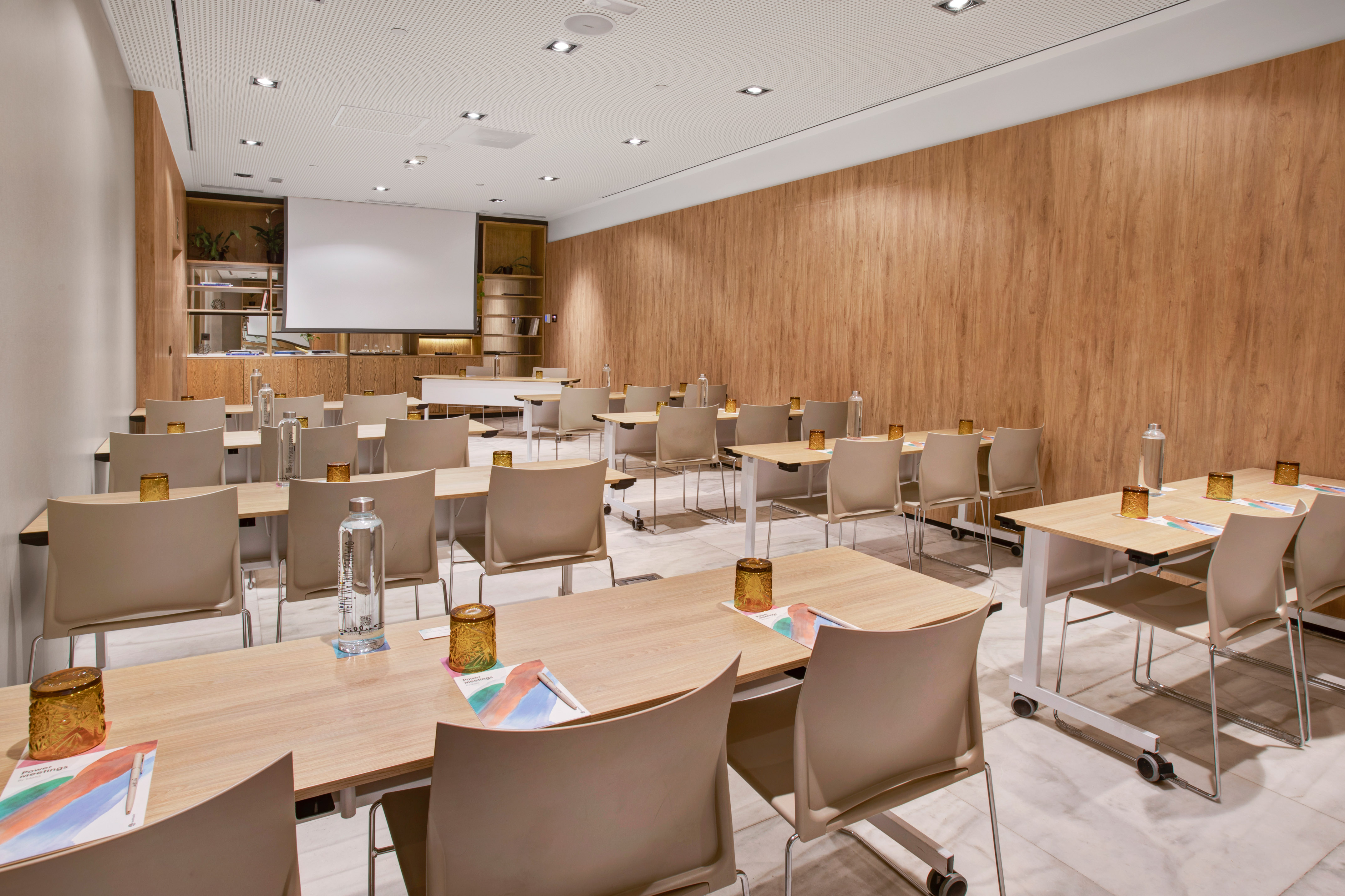 Modern meeting room with light wood tables, beige chairs, and projector screen.