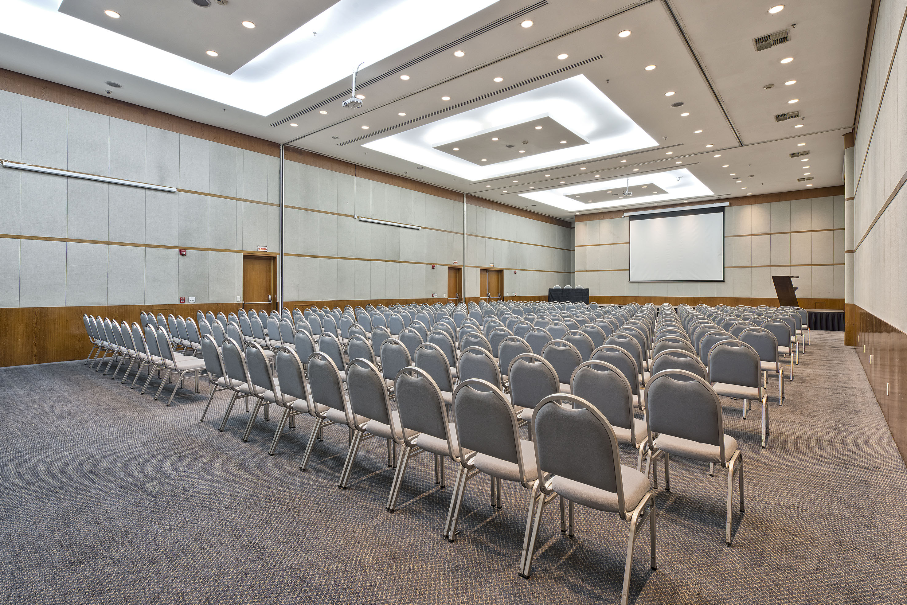Empty conference room with rows of gray chairs, projection screen, and podium.