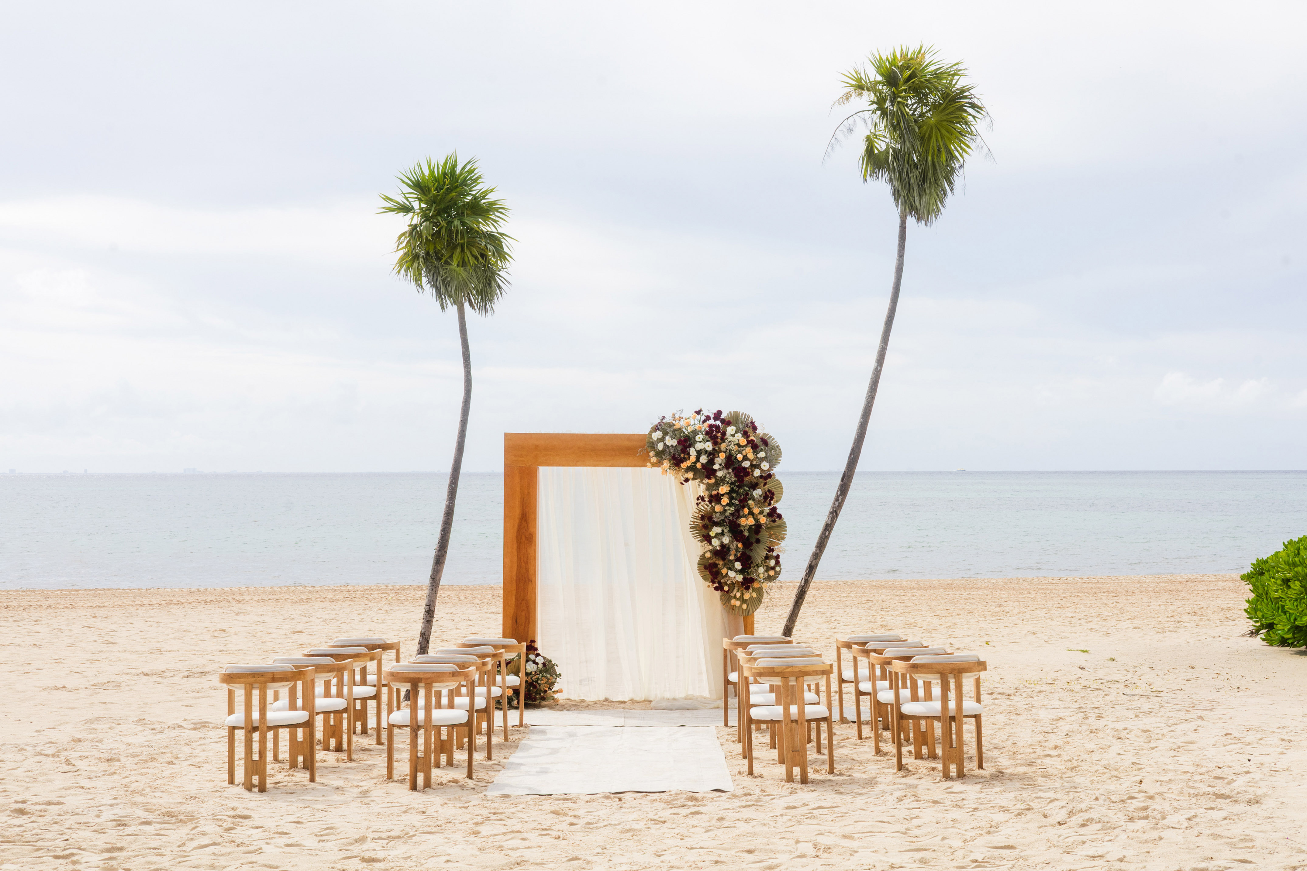 Beach wedding ceremony setup with floral arch, chairs, ocean, and palm trees.