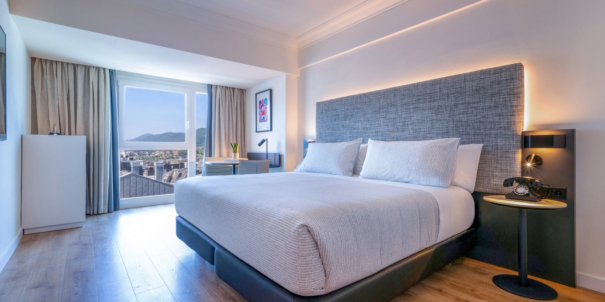 Habitación de hotel moderna con cama grande, cabecero gris y vista a la ciudad.