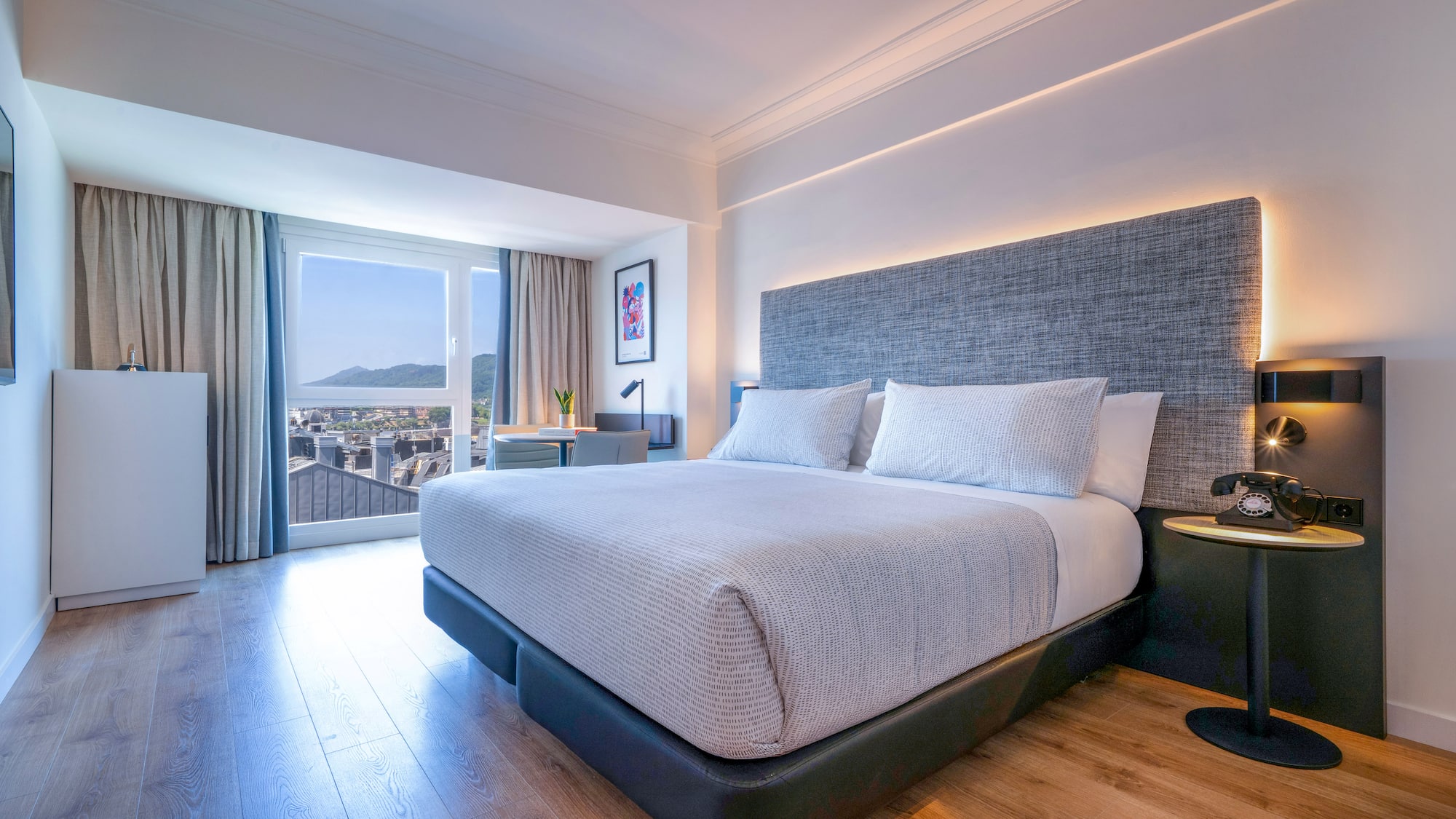 Habitación de hotel moderna con cama grande, cabecero gris y vista a la ciudad.