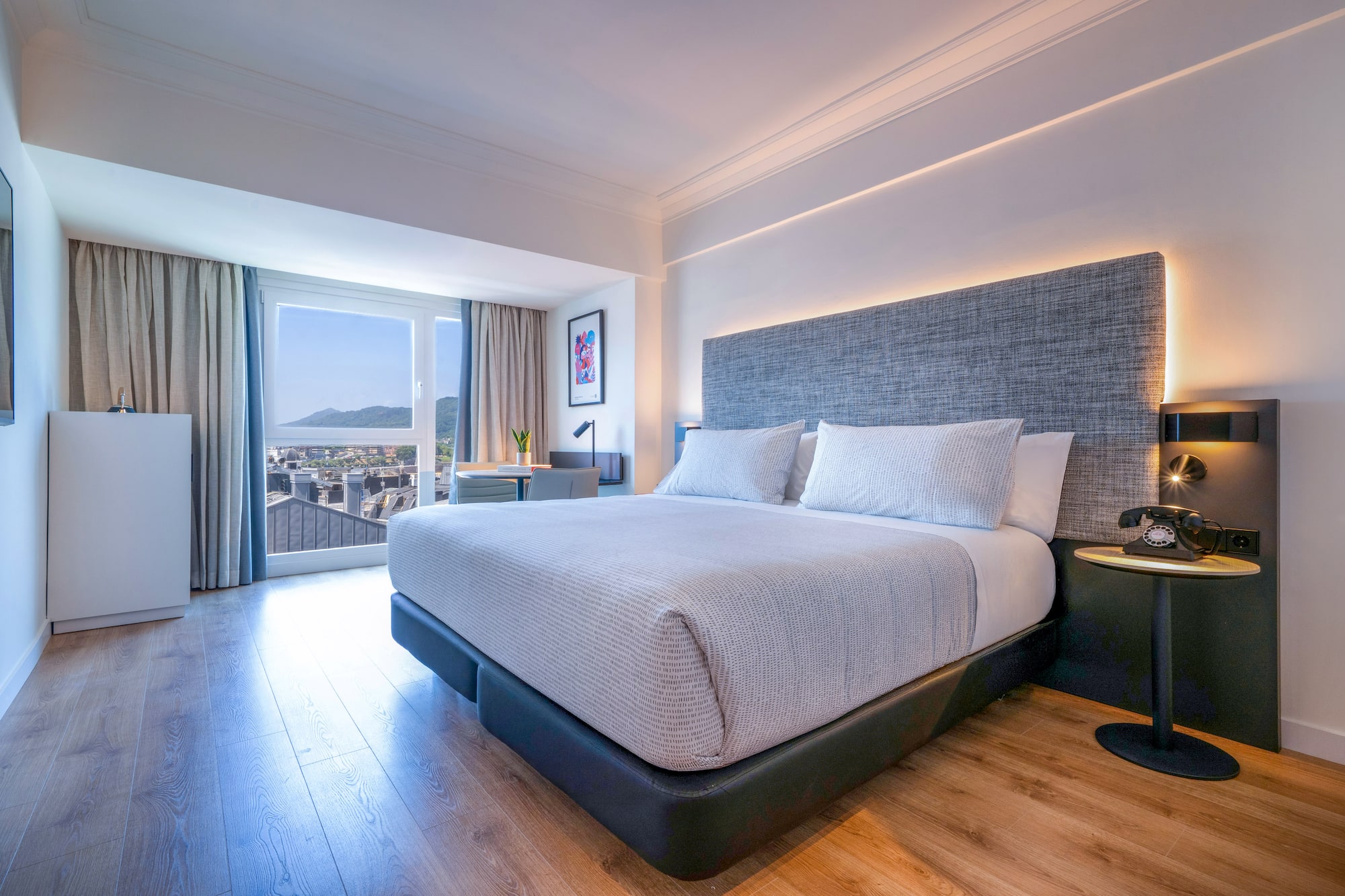 Habitación de hotel moderna con cama grande, cabecero gris y vista a la ciudad.