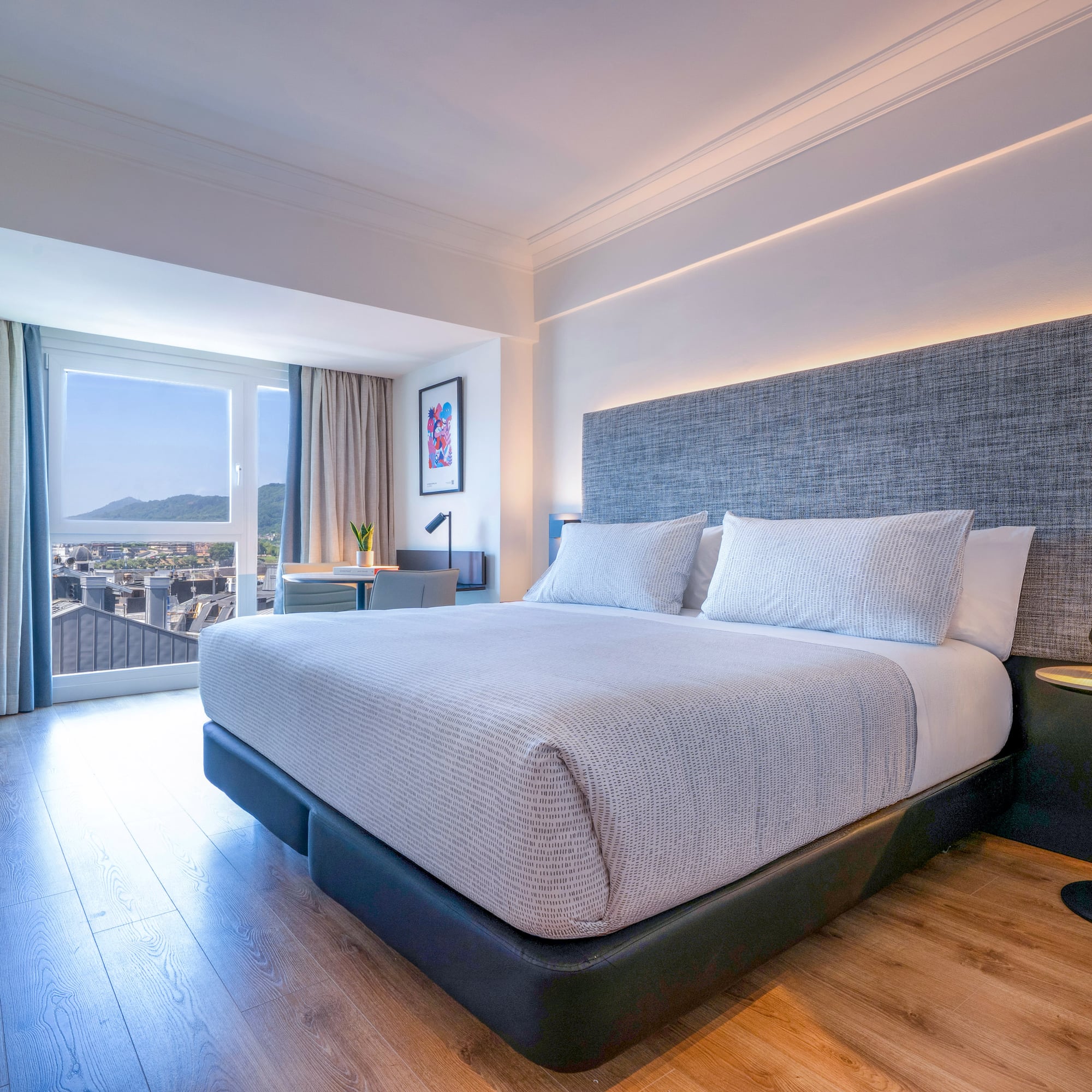 Habitación de hotel moderna con cama grande, cabecero gris y vista a la ciudad.