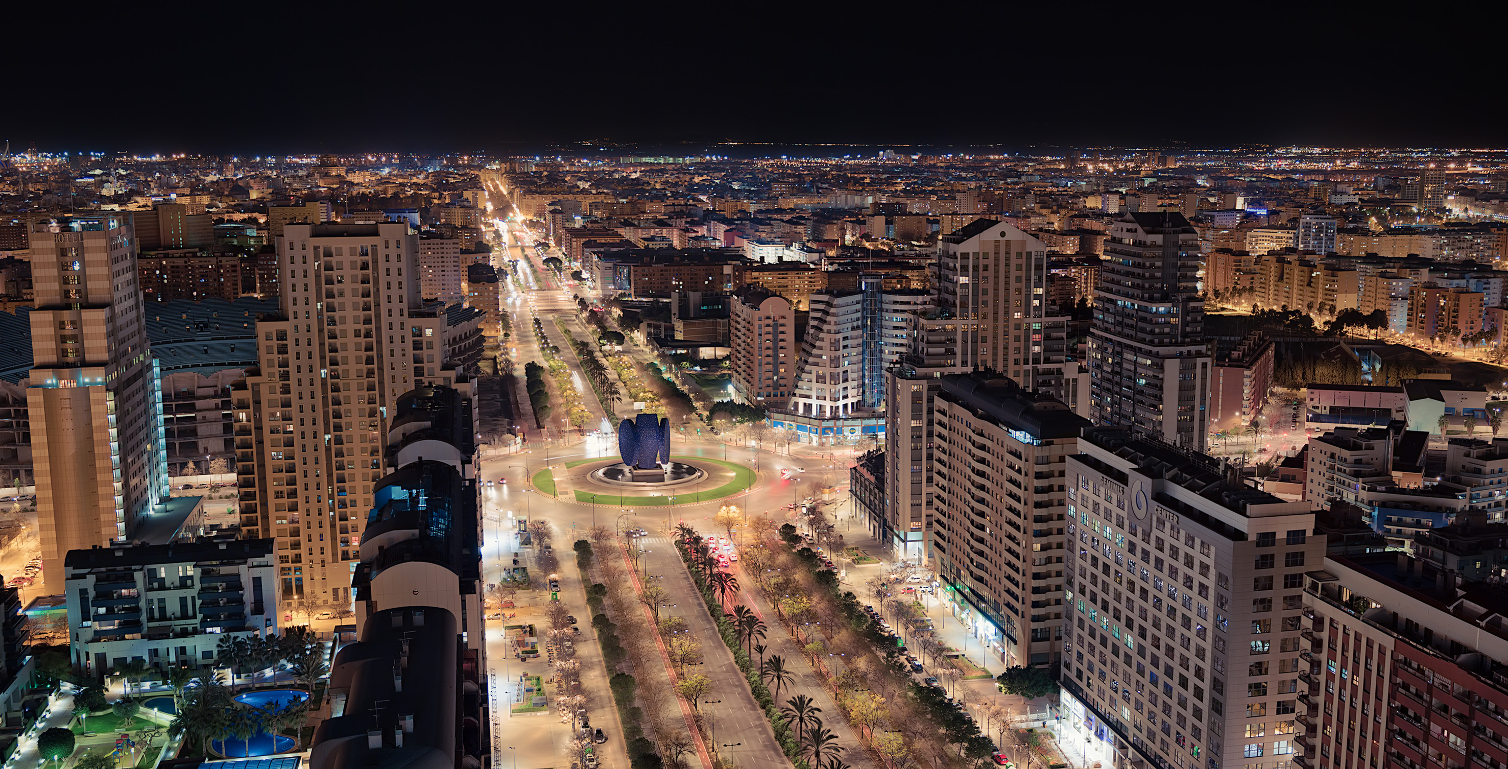 Night cityscape with illuminated streets, tall buildings, and a central roundabout.