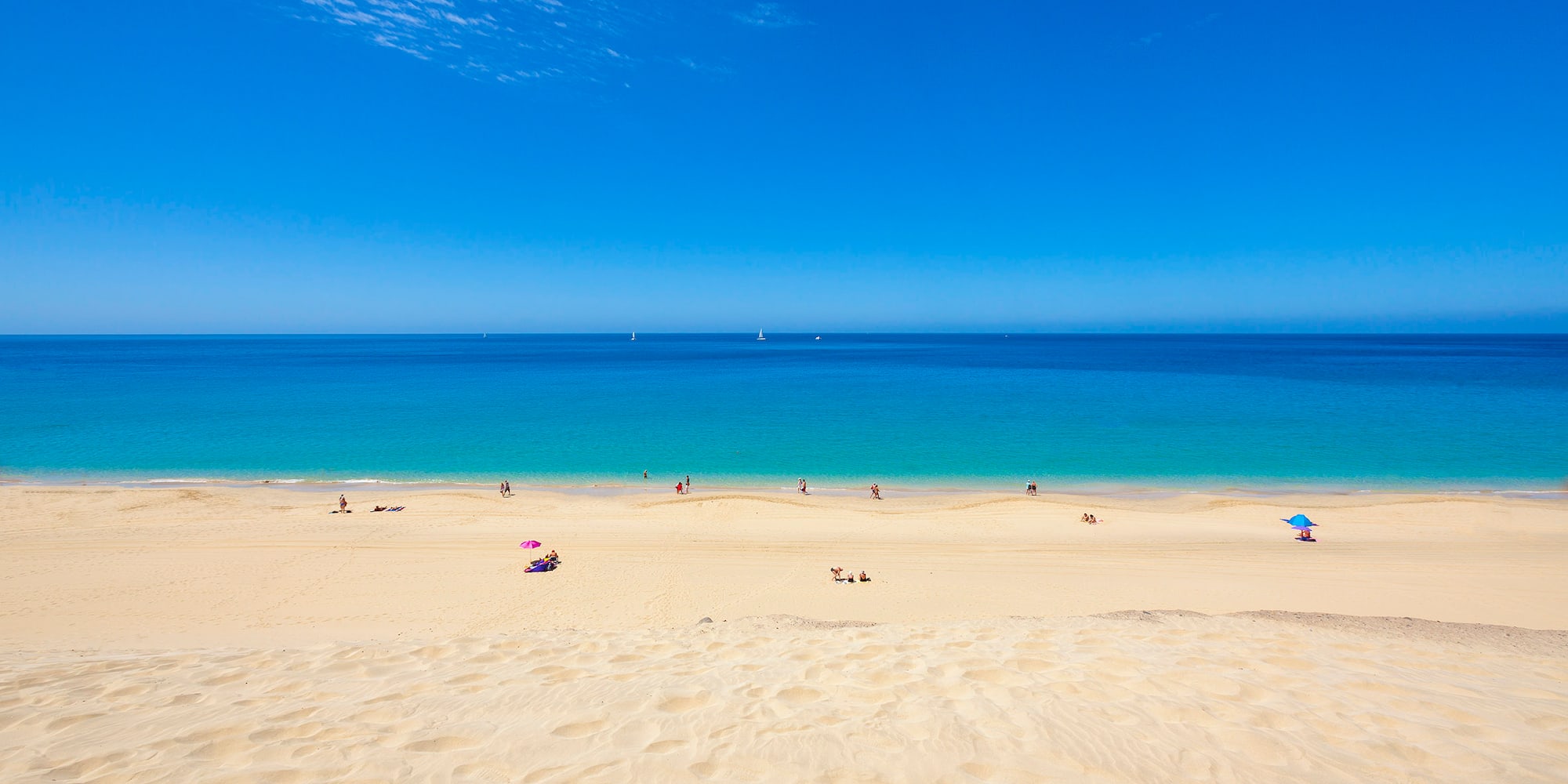 Golden sand beach, turquoise ocean, blue sky, people, distant sailboats.