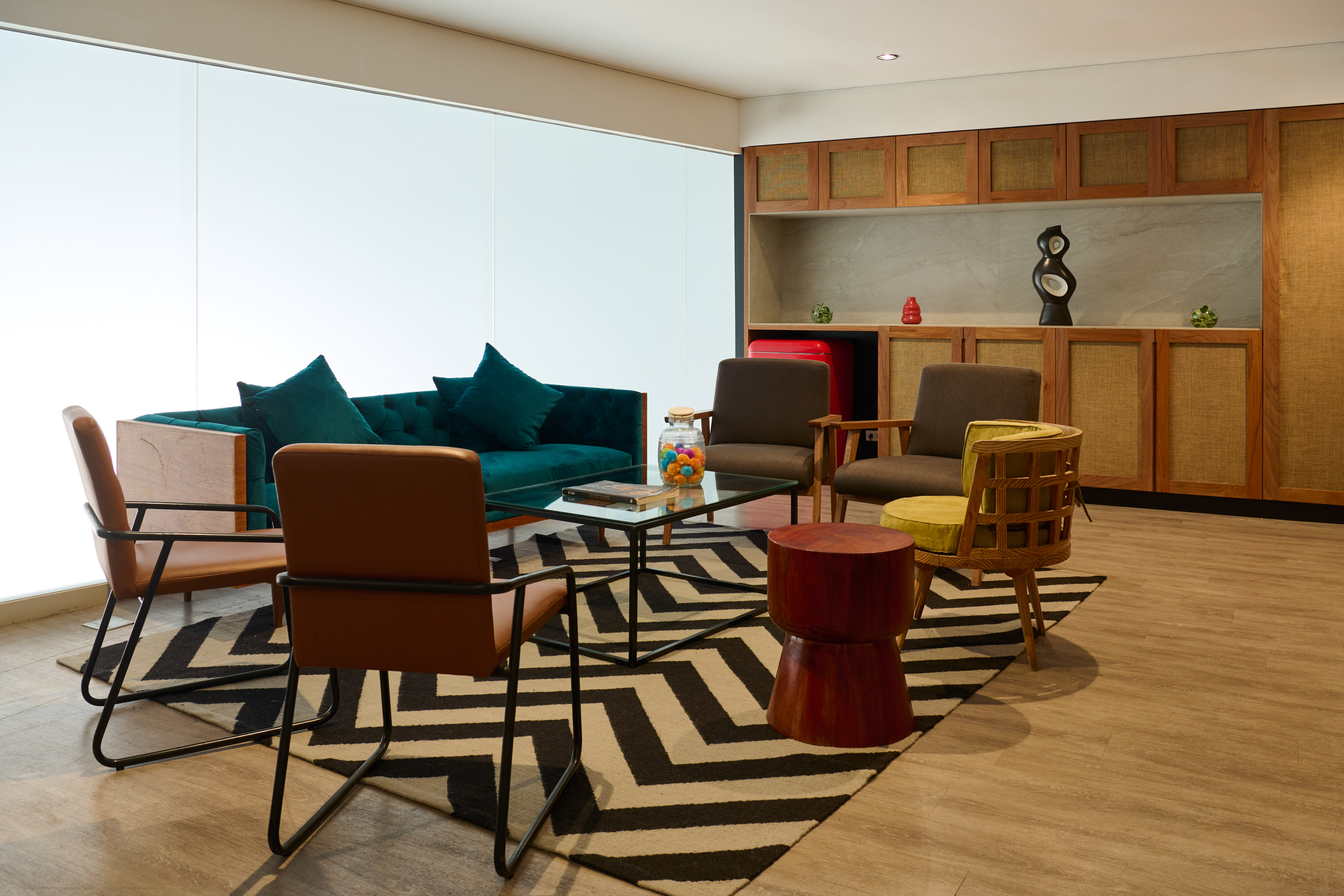 Modern lounge with teal sofa, various chairs, glass table, and geometric rug.